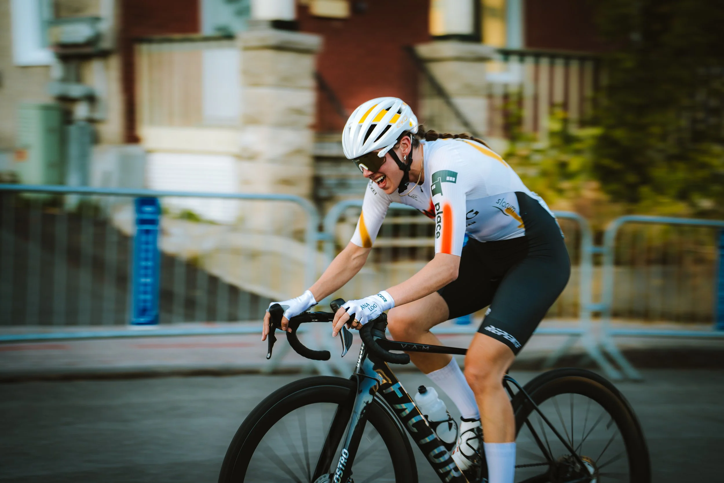 A female cyclist wearing a white and black jersey, helmet, and gloves, joyfully riding a mountain bike on a city street with a brick house in the background.