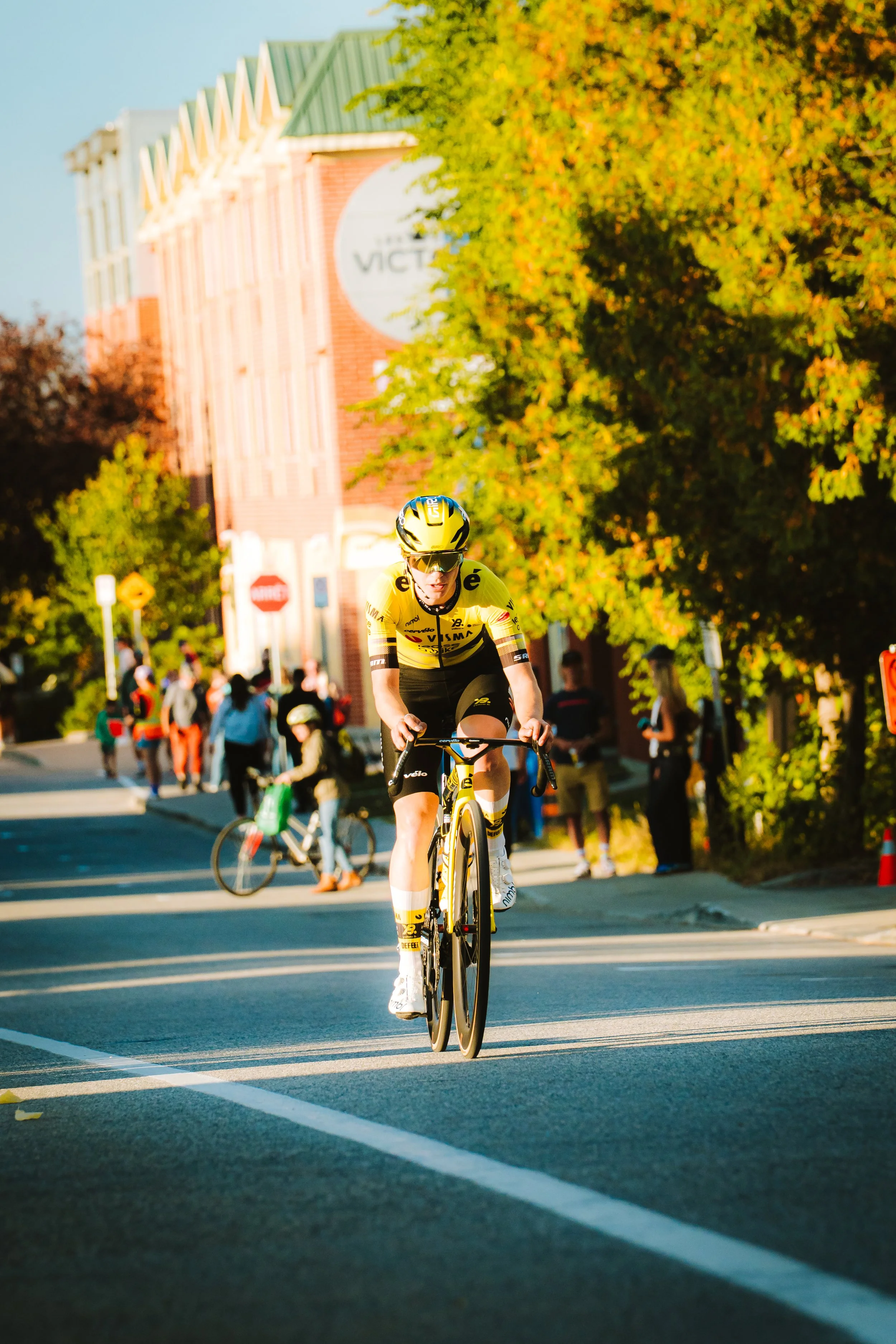 A cyclist in yellow and black riding a bike during a race on a city street with autumn trees and spectators in the background.