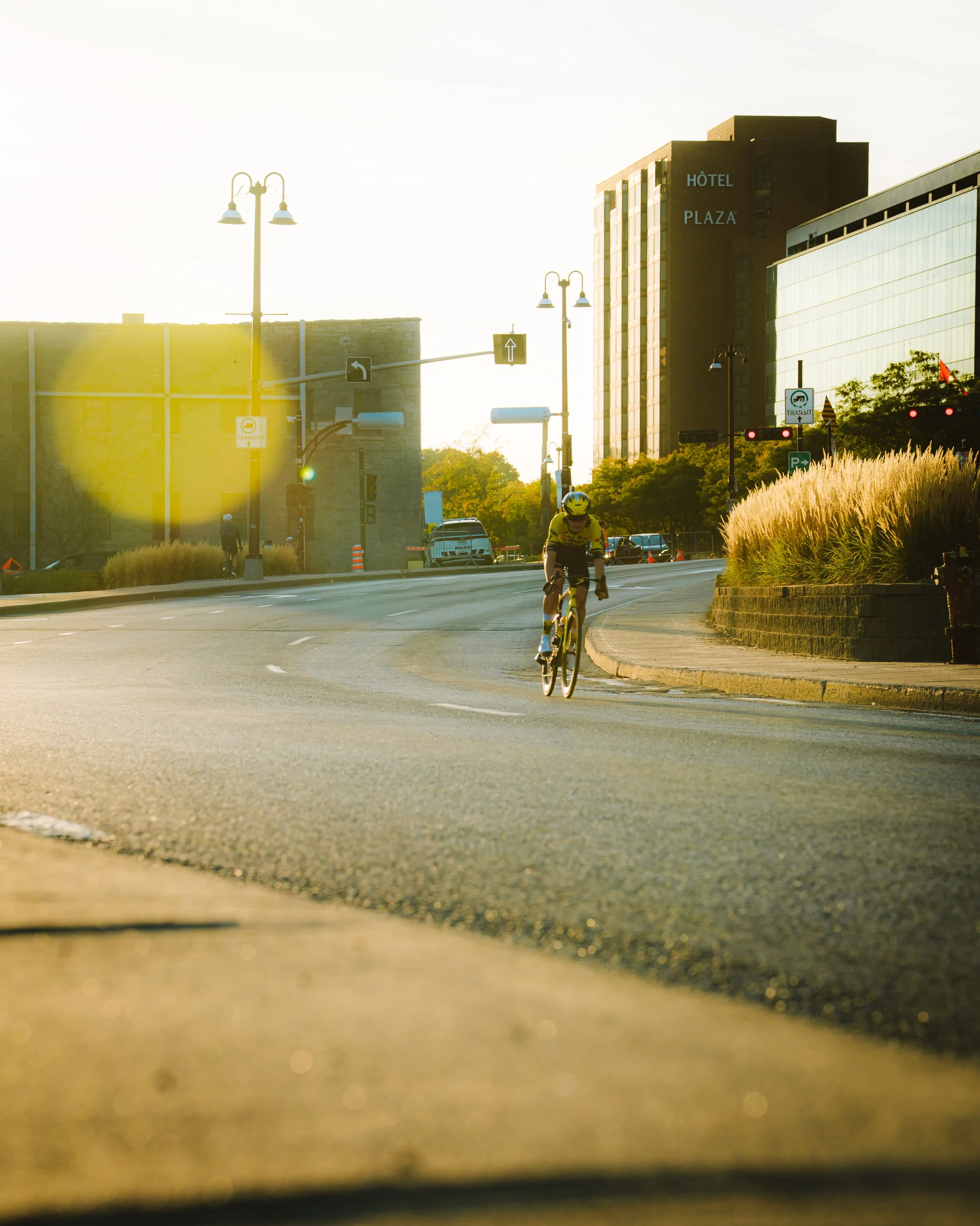 A cyclist wearing a yellow helmet and yellow shirt riding on a city street during sunrise, with modern buildings and streetlights in the background.