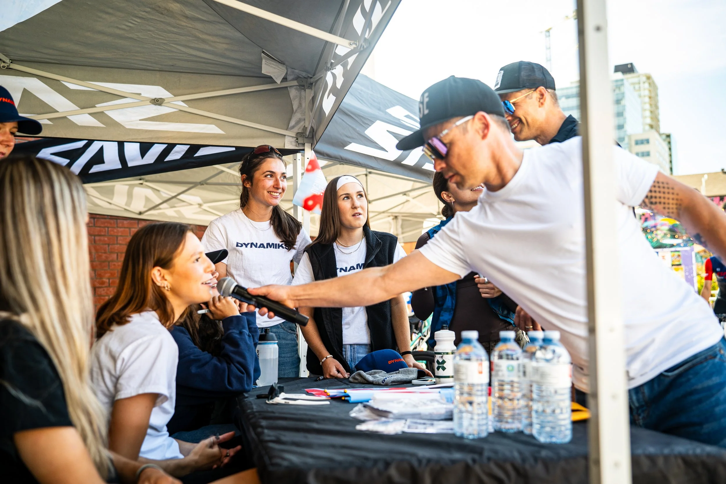 A woman sitting at a table holding a microphone is being interviewed by a man in a white shirt and cap at an outdoor event with a canopy tent and water bottles on the table.
