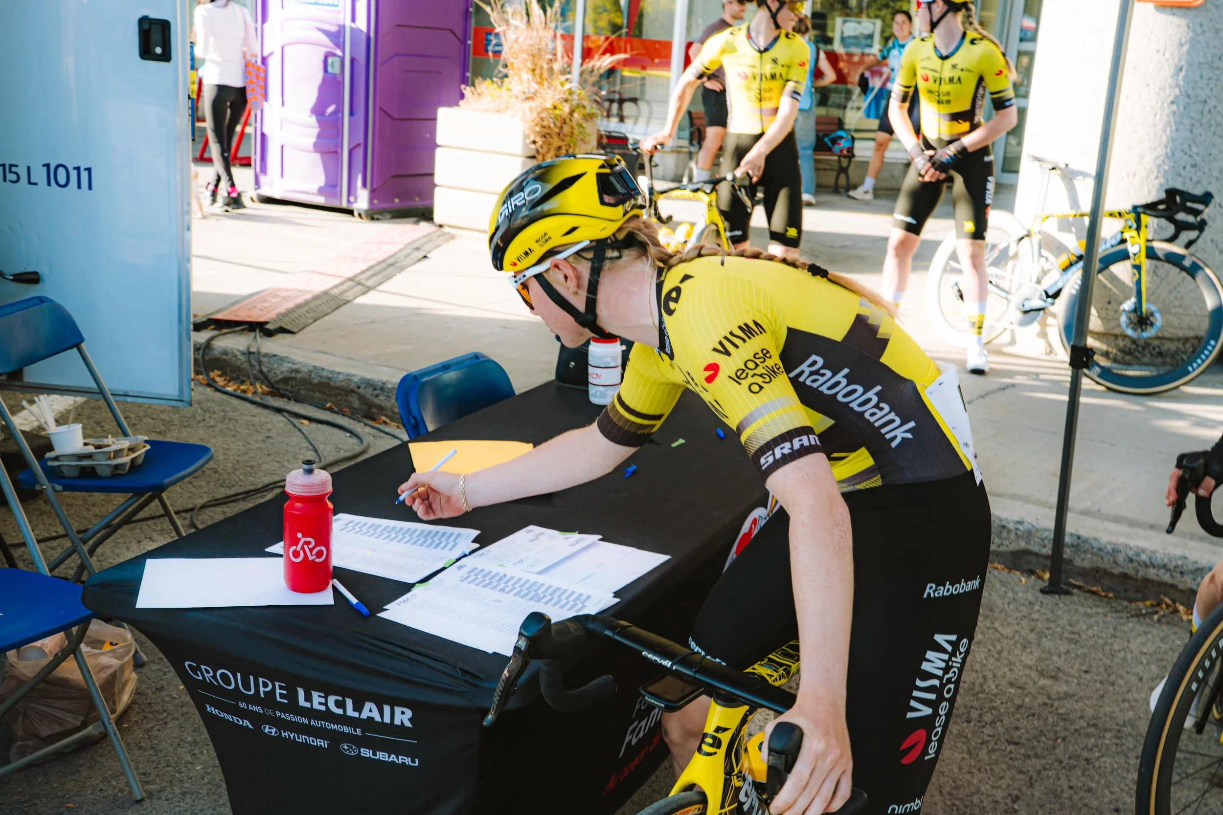 A female cyclist signing in at a registration table during a cycling event. She is wearing a yellow and black cycling jersey and helmet, with a yellow bike in front of her. Other cyclists in similar jerseys and bikes are visible in the background.