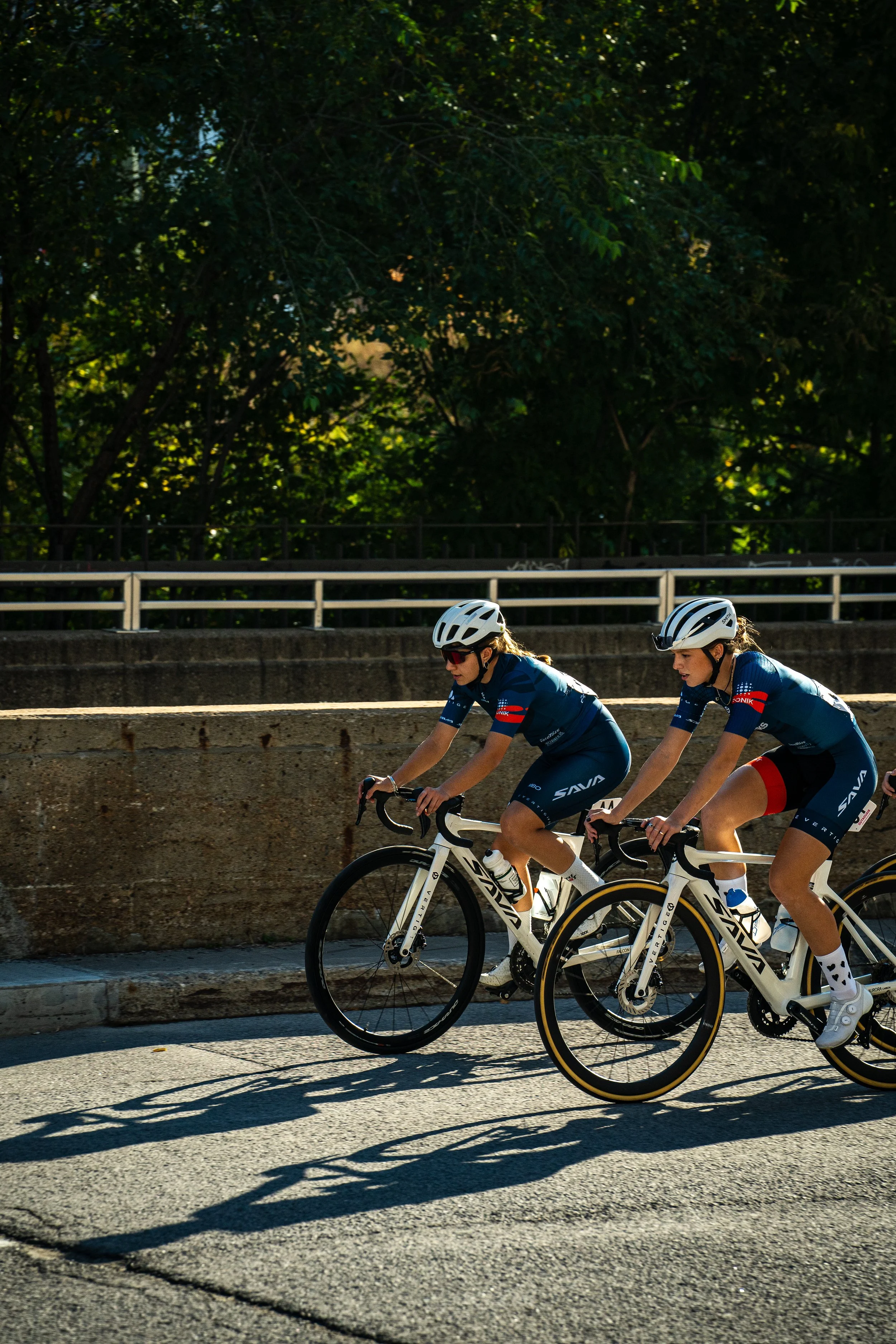 Two female cyclists riding bikes on a road during daytime with trees in the background.