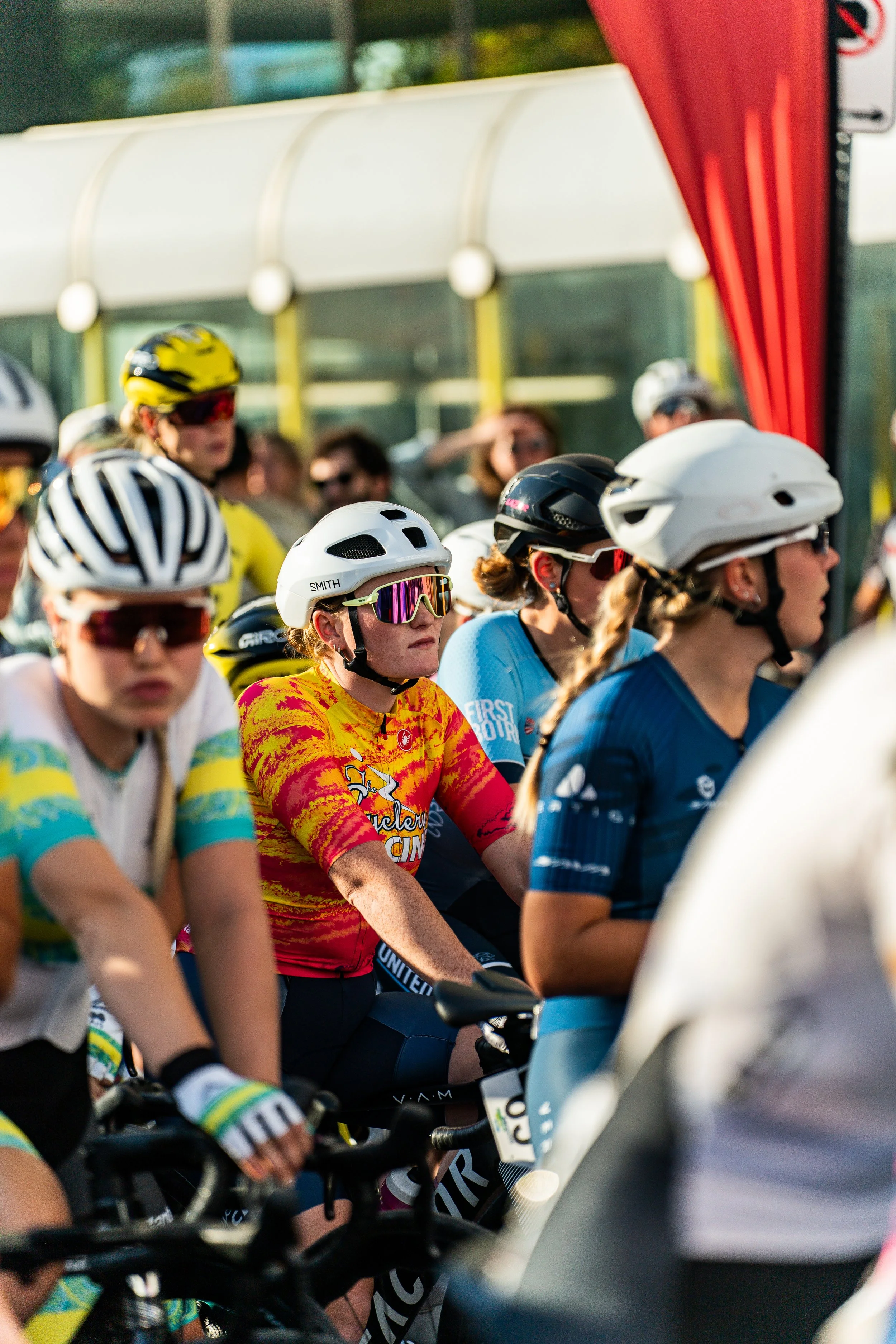 Female cyclists wearing helmets and sunglasses, gathered at the starting line of a cycling race, with a red banner in the background.