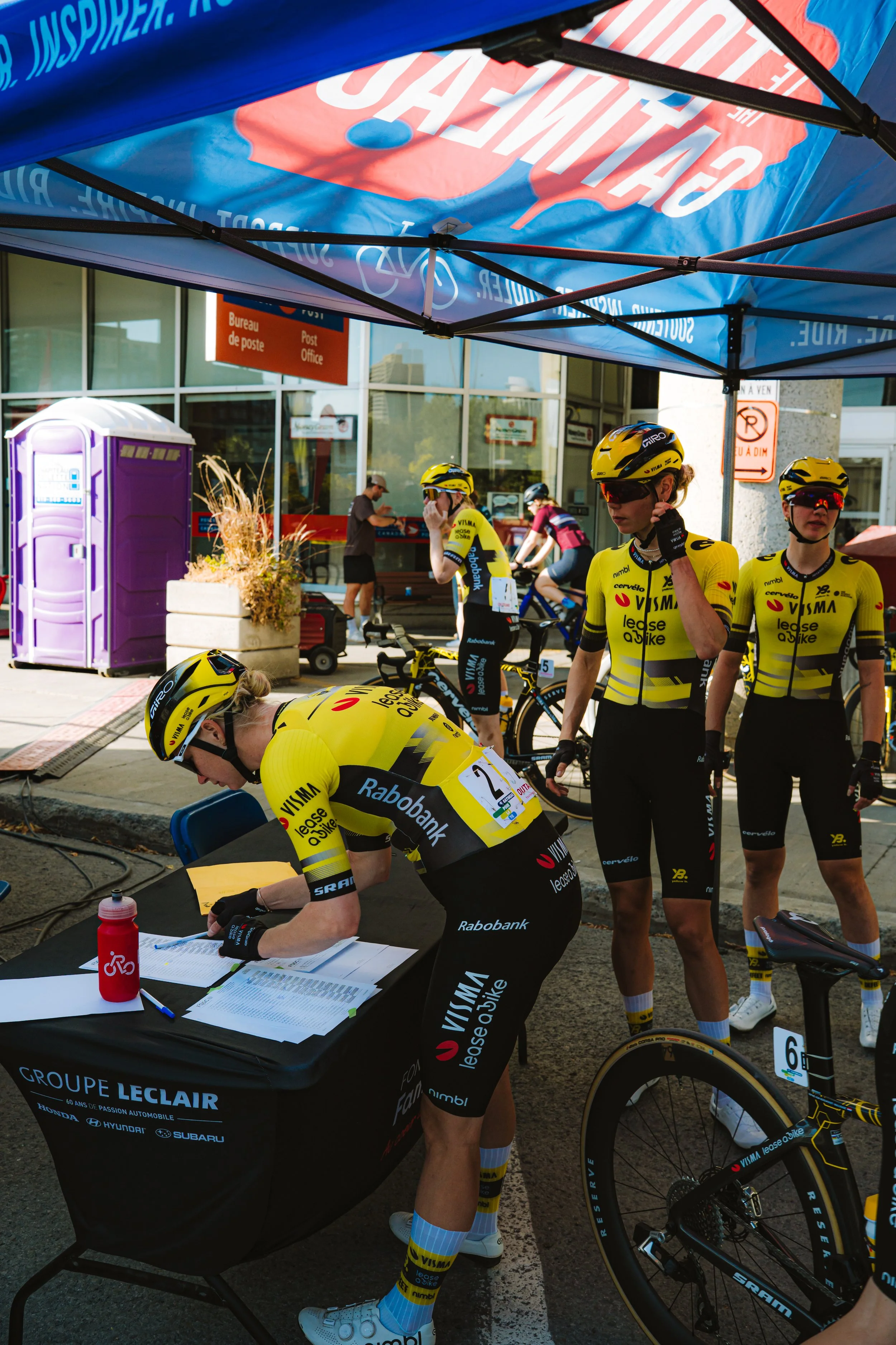 Cyclists in yellow team jerseys at a registration table during a race, with a tent overhead and a post office building in the background.