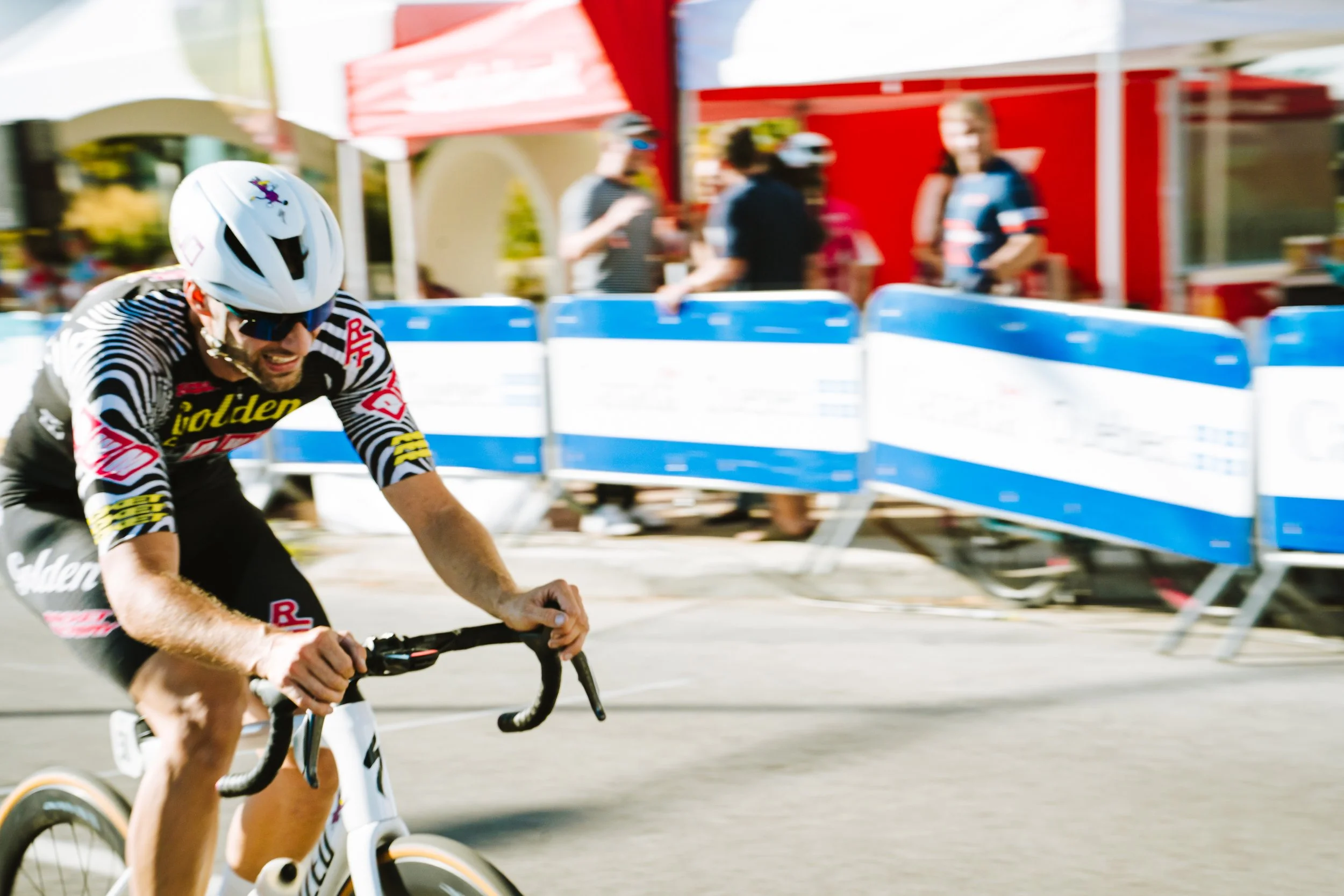 A male cyclist in a black and white striped jersey with yellow and red accents, wearing a white helmet and sunglasses, is racing on a road during a cycling event with blue barriers and tents in the background.