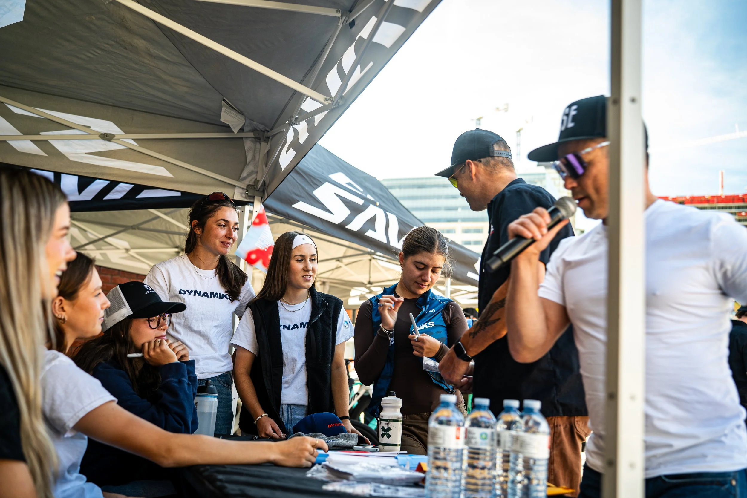 Group of young women and two men gathered under a canopy at an outdoor event, with one man speaking into a microphone and the women listening. There are water bottles on the table.