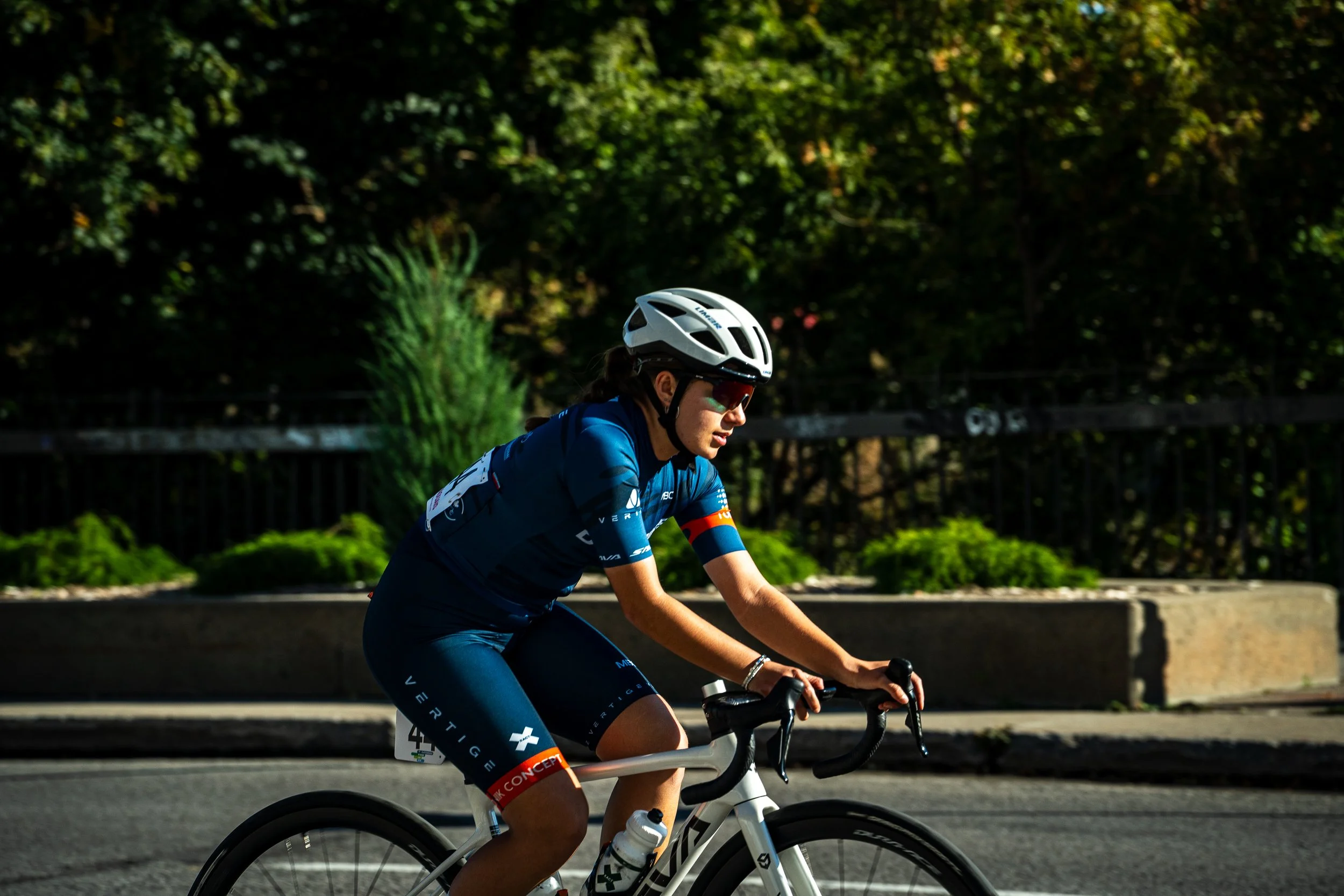 Female cyclist wearing a white helmet, sunglasses, and a blue cycling jersey riding on a paved road with trees and green shrubs in the background.