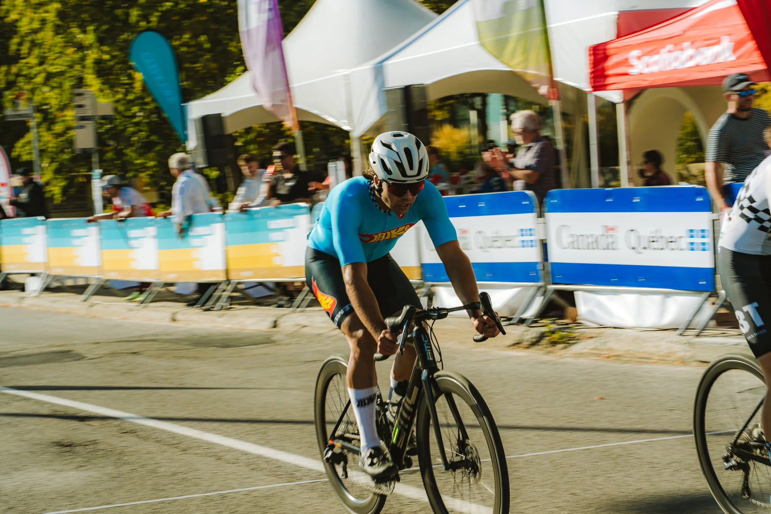 Cyclist racing in a race, wearing a blue jersey, black shorts, helmet, and sunglasses, on a paved street with spectators and banners in the background.