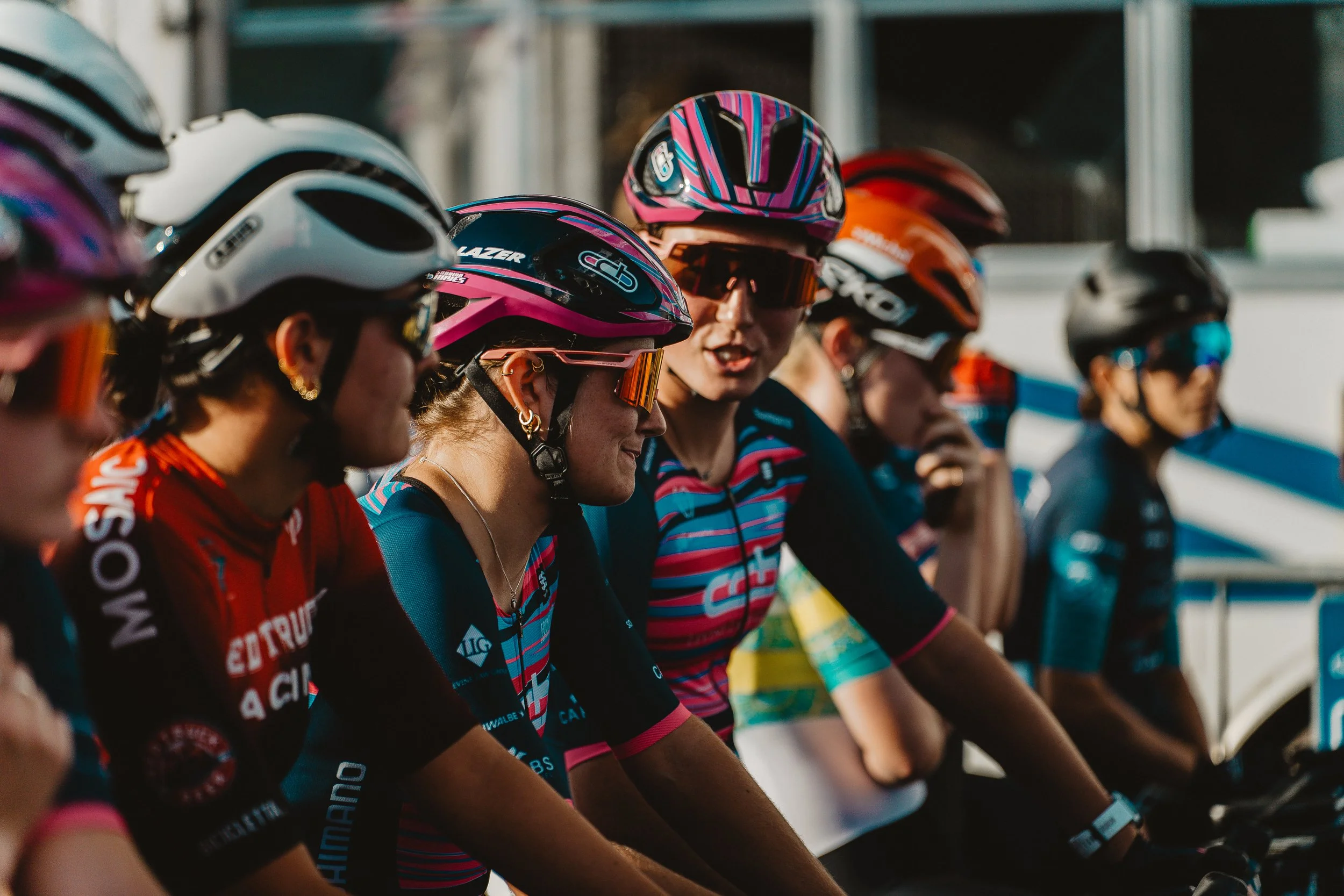 Group of female cyclists wearing helmets and colorful jerseys, ready for a race.
