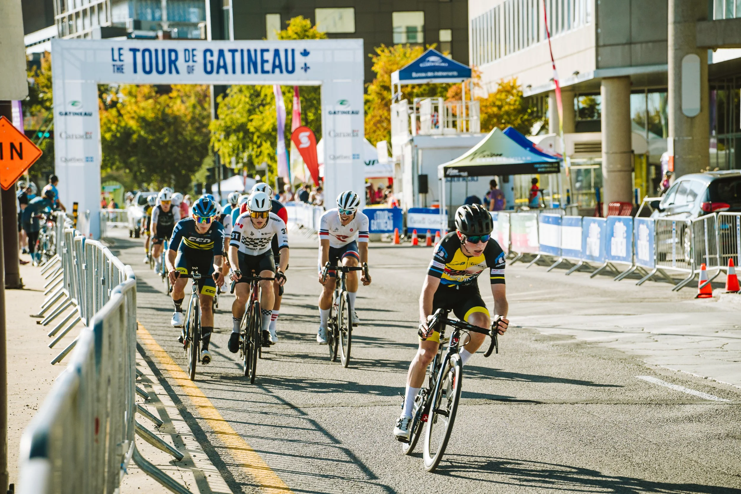 Cyclists competing in a race on a city street marked with barriers, with a banner reading 'Le Tour de Gatineau' overhead, and spectators watching from the sidelines.