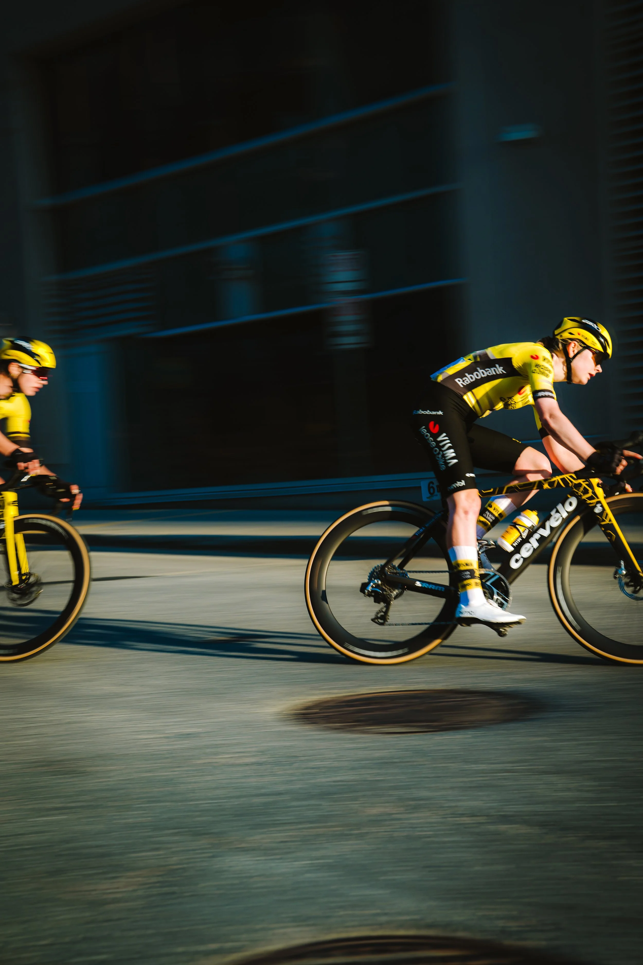 Cyclists racing on a city street, with one cyclist in a yellow and black outfit riding a black and yellow bike, and another cyclist partially visible on the left.