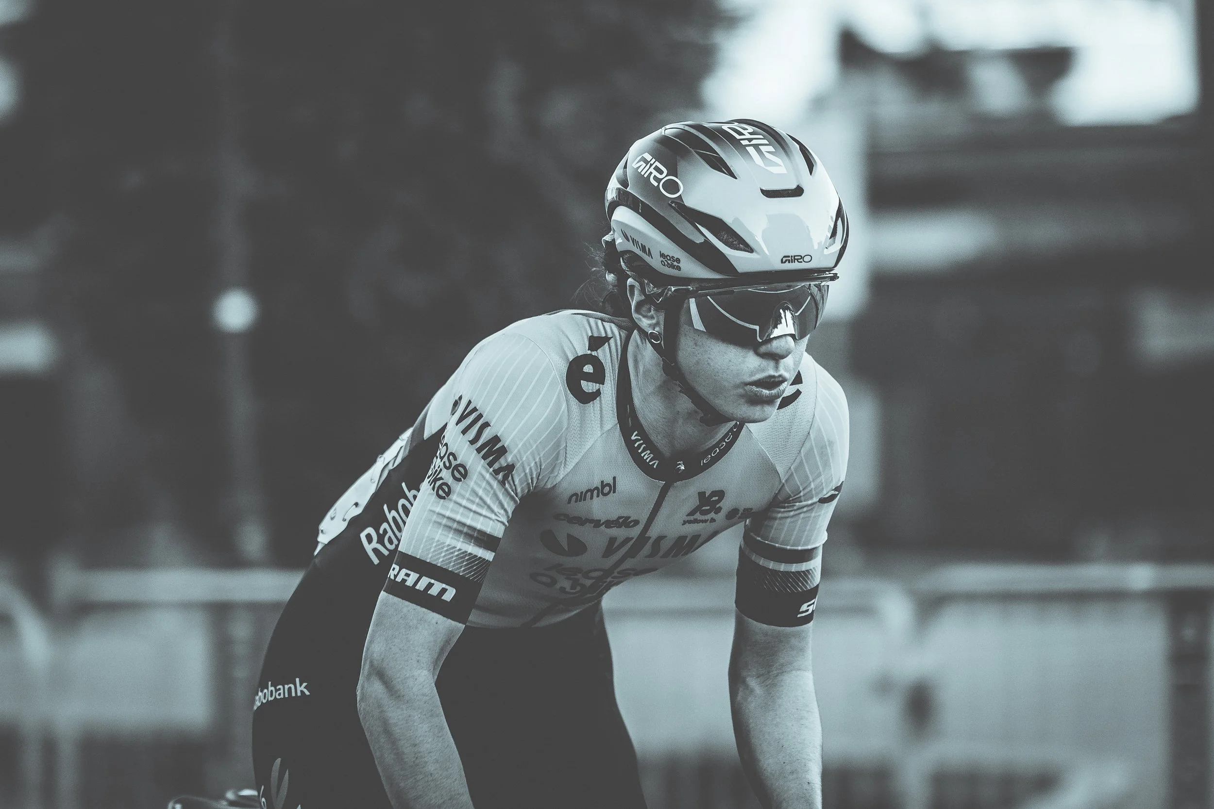 A female cyclist in a racing uniform and helmet, wearing sunglasses, preparing to race on a road.