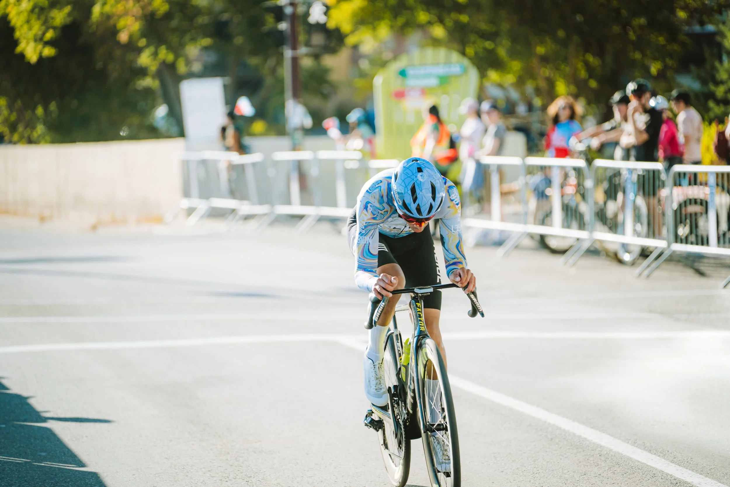 A cyclist in a blue helmet and colorful jersey racing on a city street, with spectators and bicycles lined up behind barriers in the background.