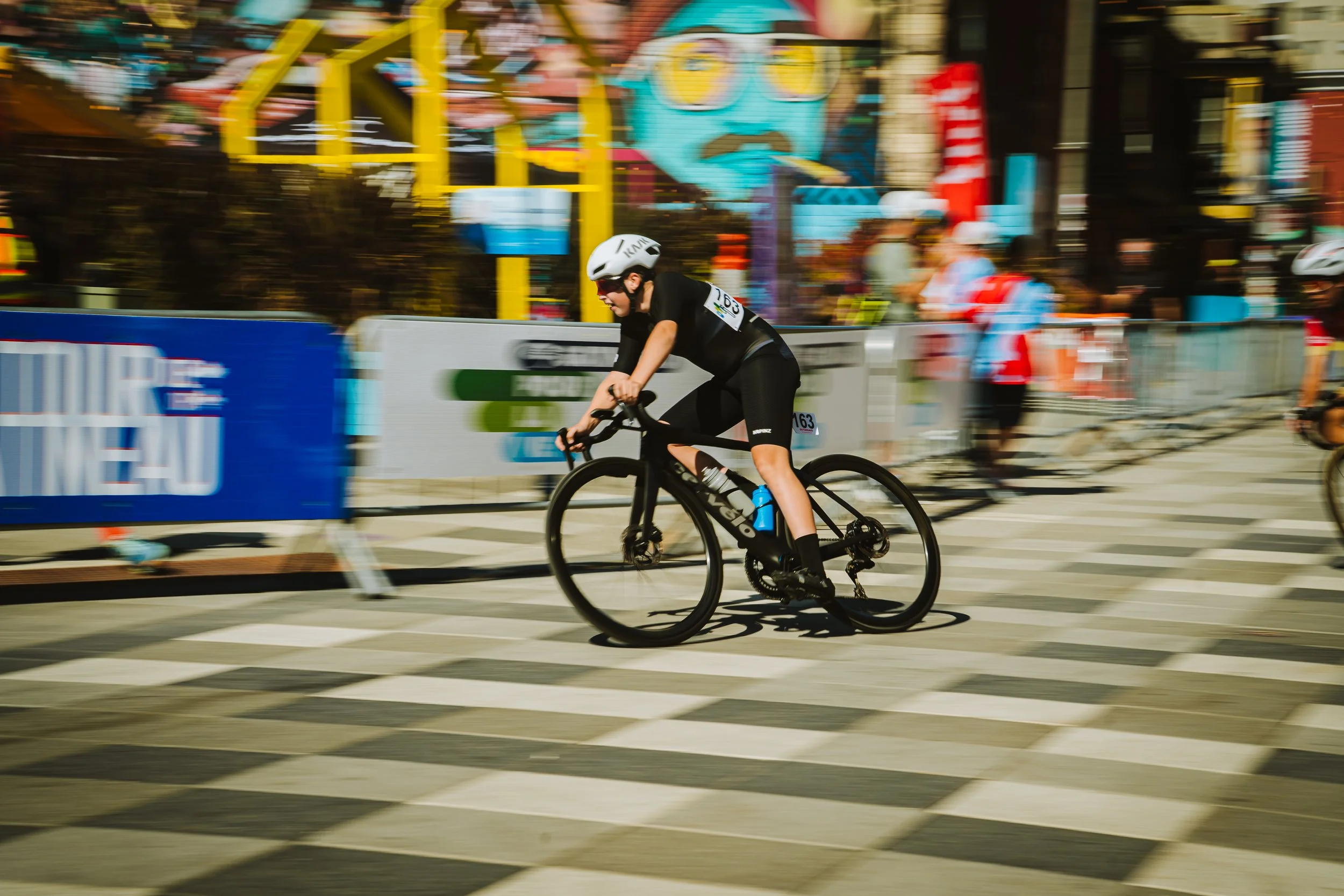 A cyclist wearing a helmet and black sports attire riding a black bicycle on a city street during a race, with colorful signs, artwork, and spectators in the background.