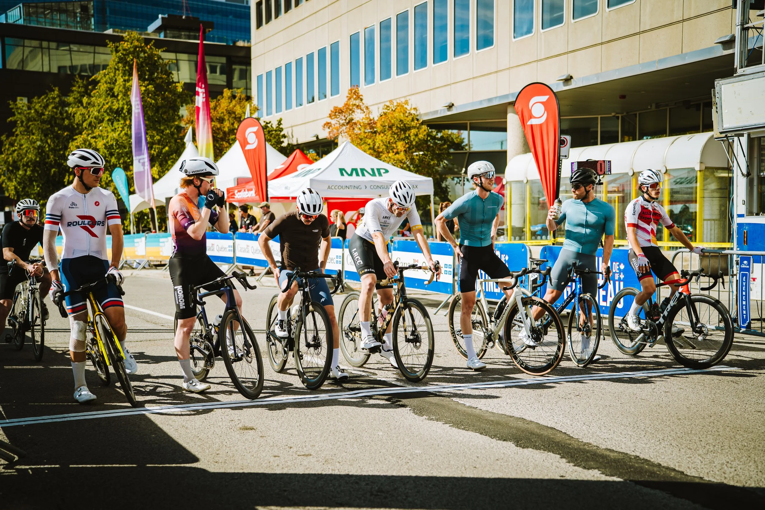 Cyclists gathered at the start line of a race in an urban setting on a sunny day, with tents and flags in the background.