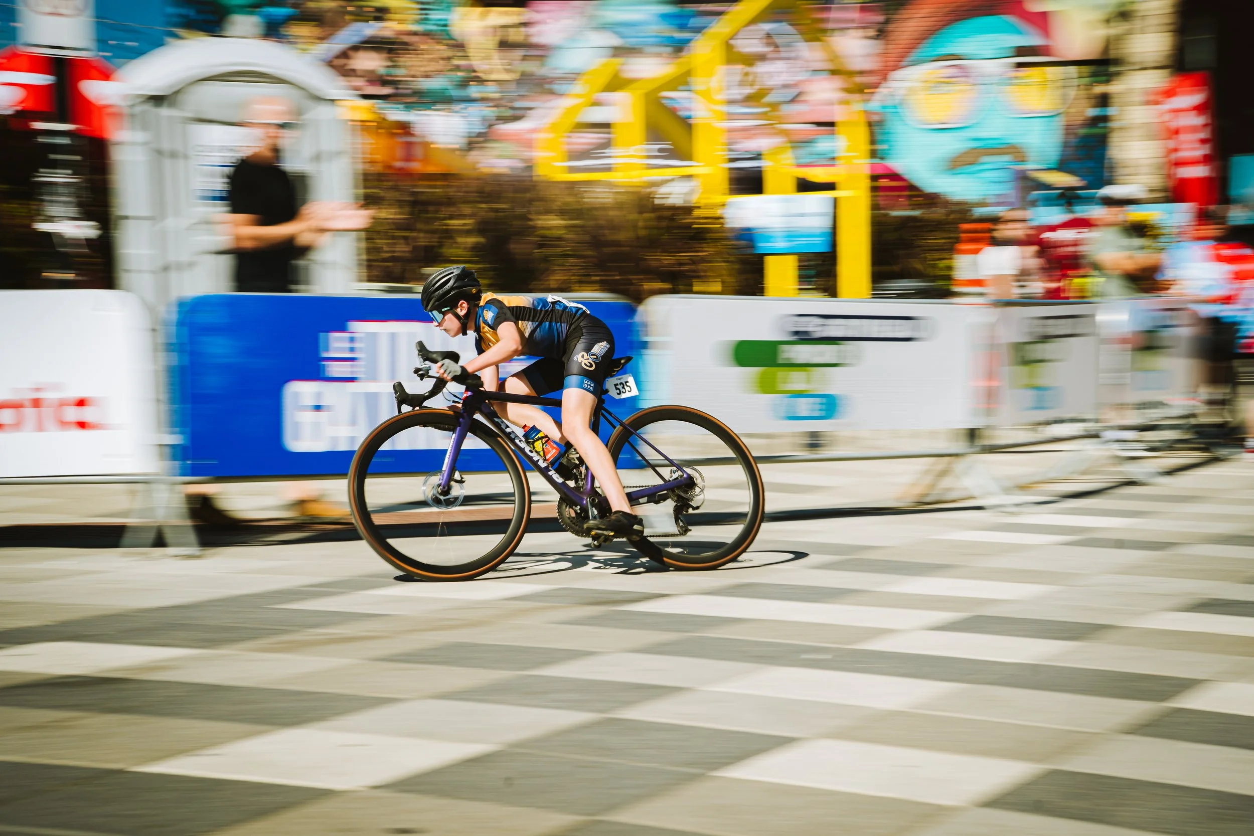 A cyclist wearing a black helmet and blue jersey riding a black and purple bike during a race, capturing the motion blur of the event background.