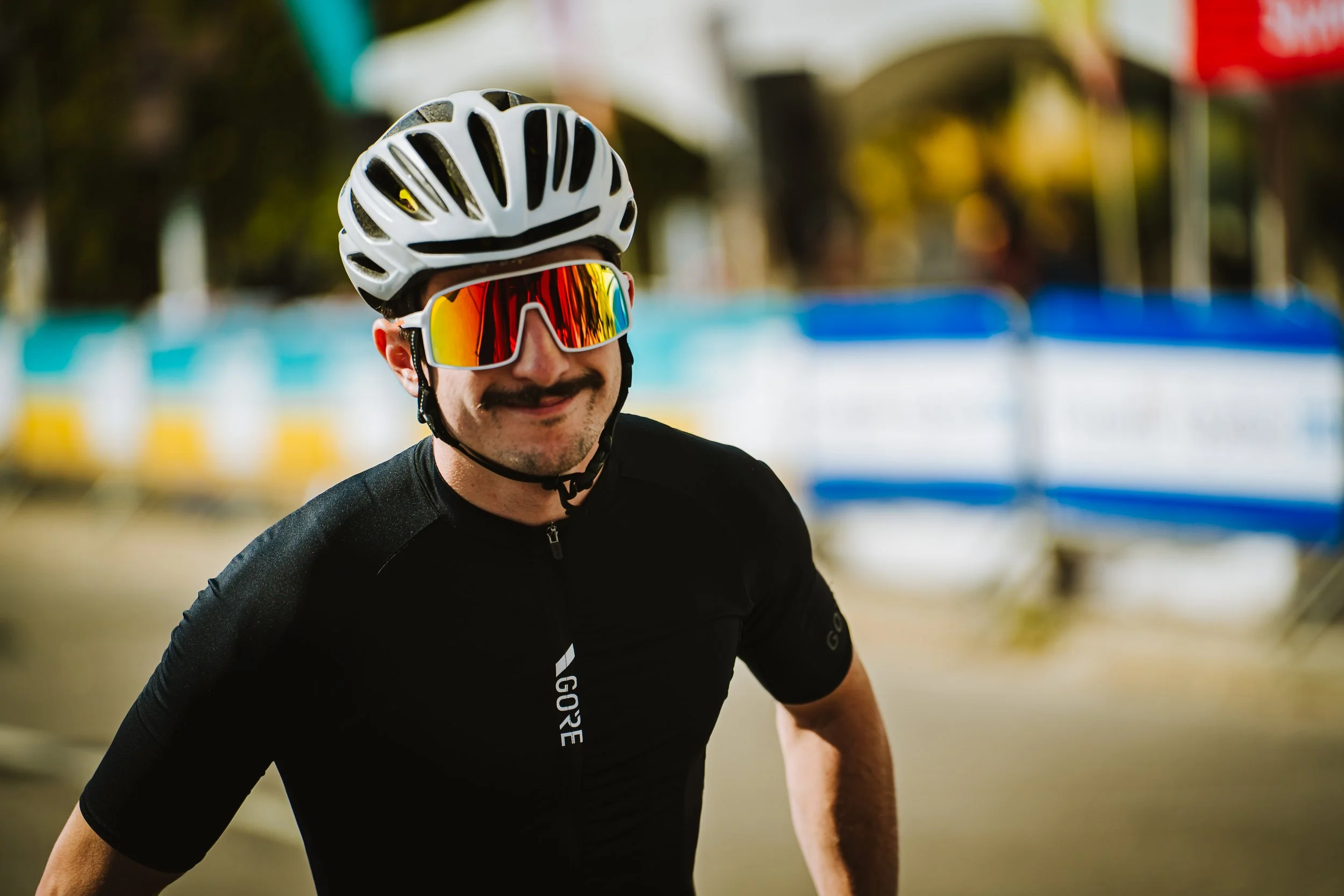 A man wearing a white bicycle helmet and colorful UV-protection sunglasses, smiling, dressed in a black cycling jersey, in an outdoor setting with barriers and blurred background.