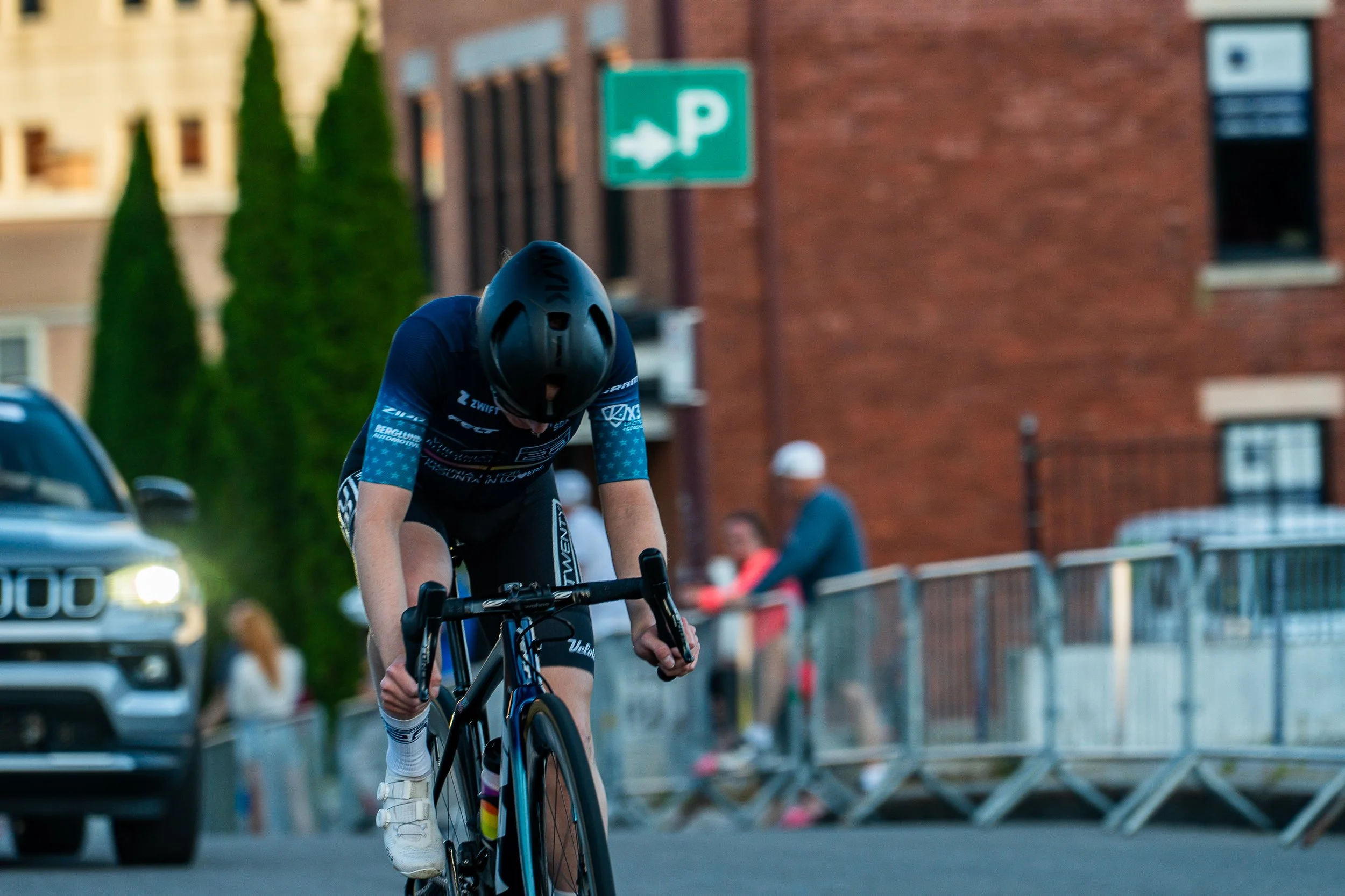 A cyclist wearing a black helmet and a blue and black cycling outfit riding a black and blue road bike on a city street, with a brick building, a green parking sign, and onlookers in the background.