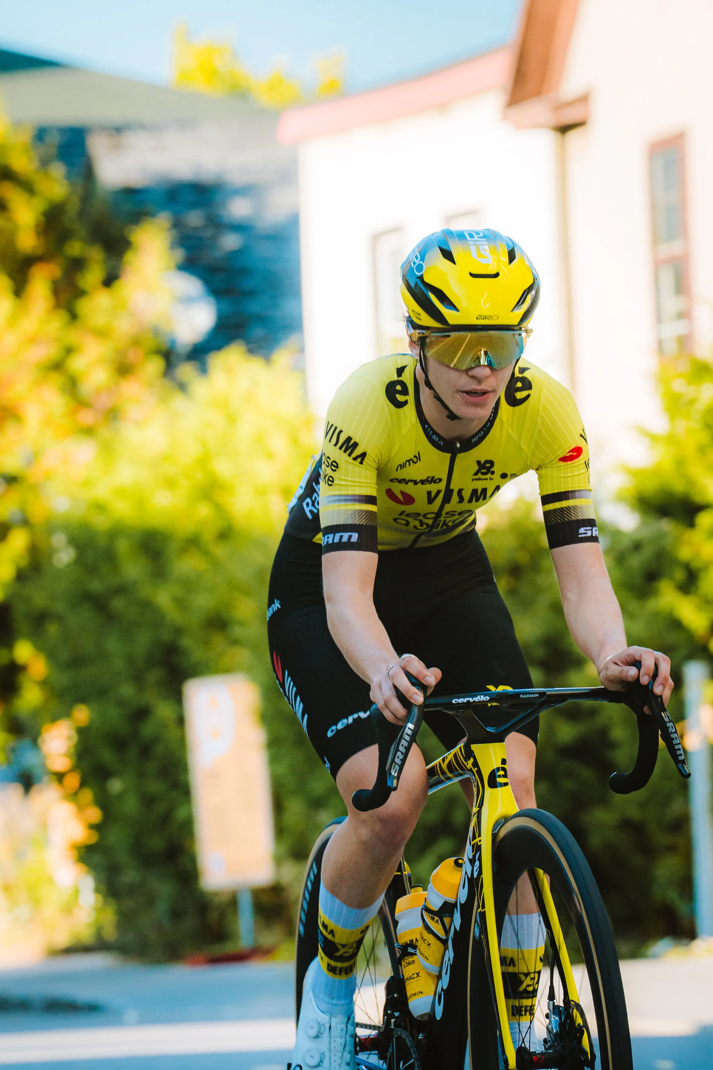 A cyclist riding a yellow and black road bike, wearing a yellow helmet, yellow riding jersey, and sunglasses, on a street with greenery and a house in the background.