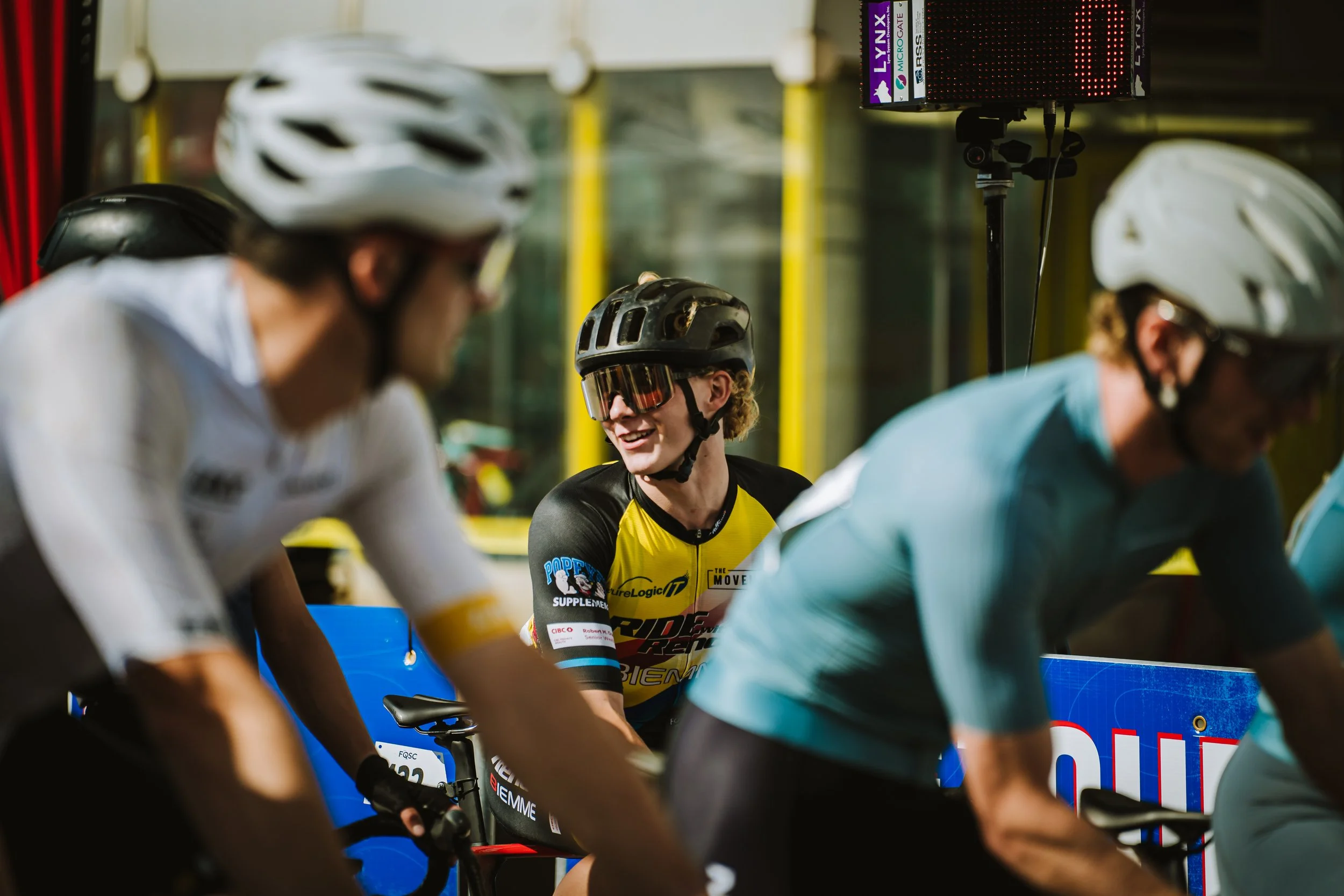 Cyclists wearing helmets and sports gear at the start of a race, with one smiling cyclist at the center, surrounded by others on bikes.