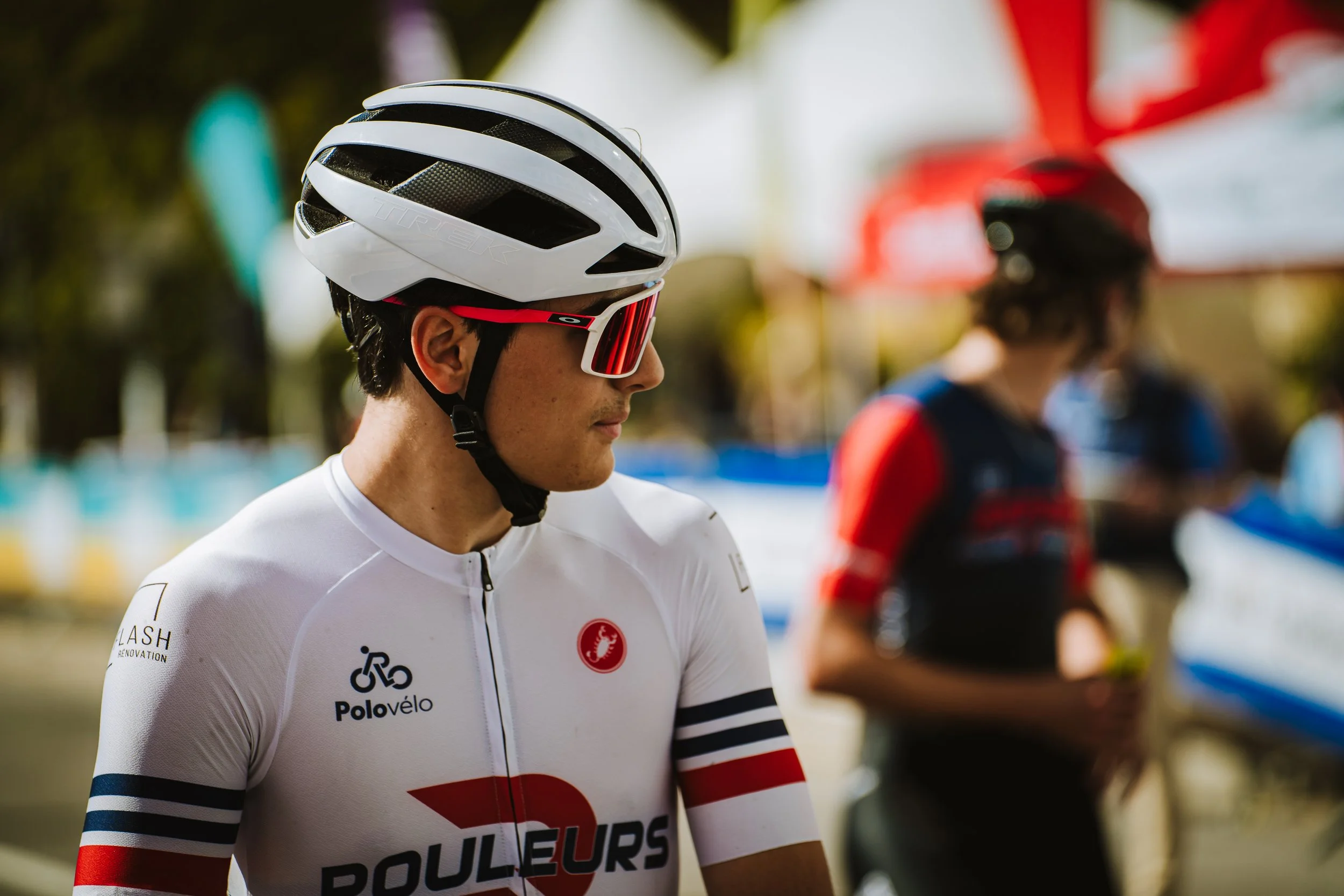 A male cyclist wearing a white helmet, red sunglasses, and a white cycling jersey with red, black, and blue accents. He is outdoors with a blurred background. Another cyclist in a black and red jersey is also visible in the background.