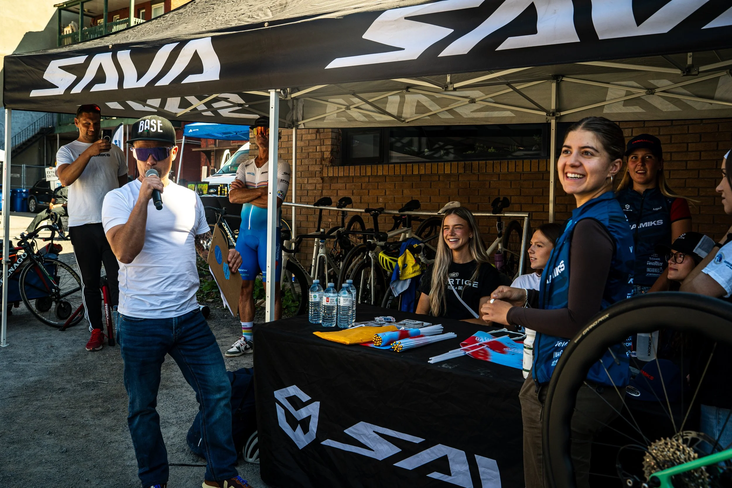 A group of people, including a man with a cap and sunglasses speaking into a microphone, and women wearing biking gear, at a table under a tent with the word 'SAVA' on it, at a cycling event with bicycles in the background.