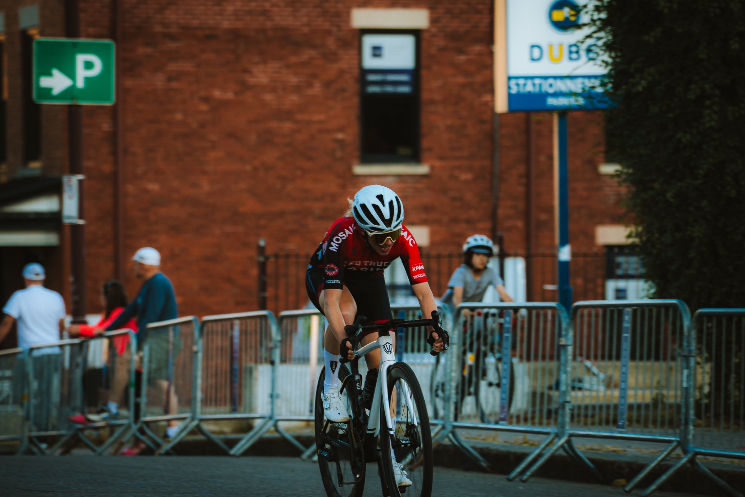 Cyclist in red and black uniform wearing a white helmet riding a bike on a city street, with barriers and onlookers in the background.