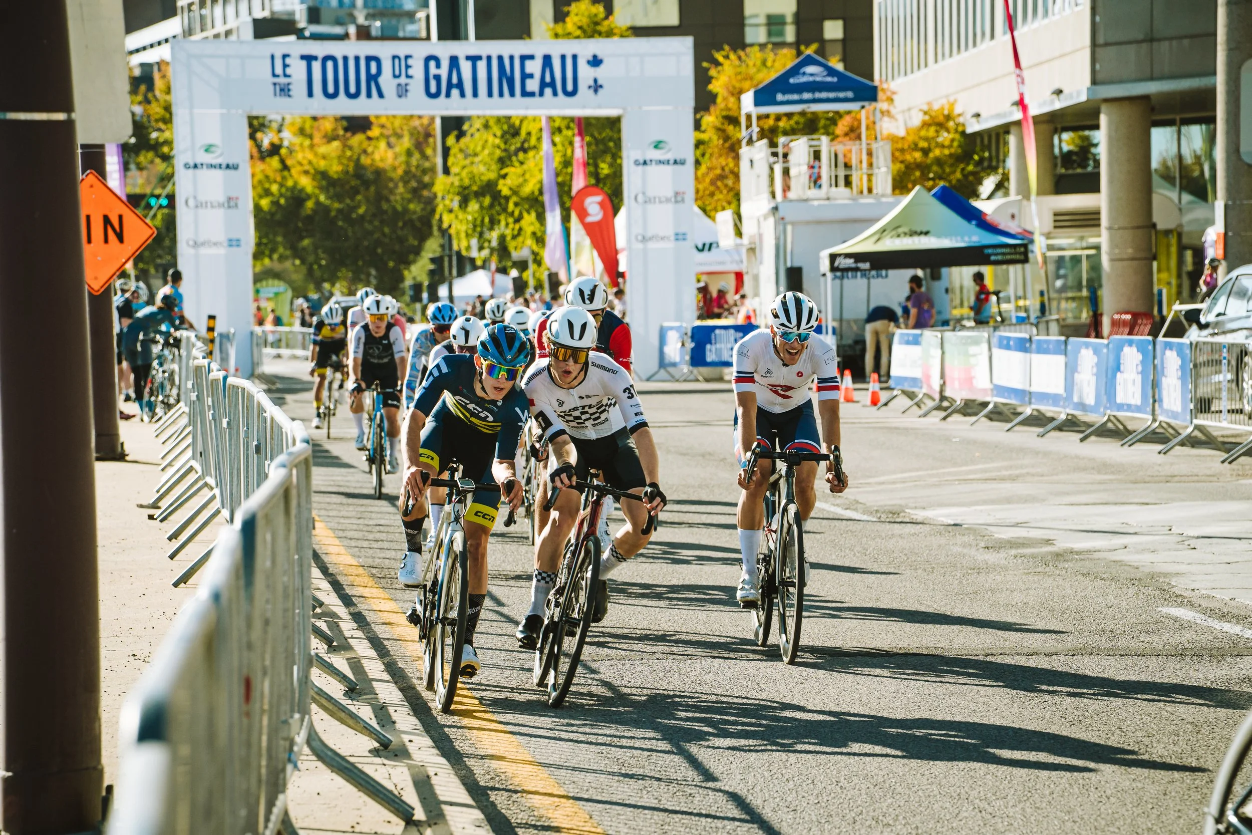 Cyclists competing in a race while riding on a city street with barriers, trees, and event signage overhead that reads 'Le Tour de Gatineau'