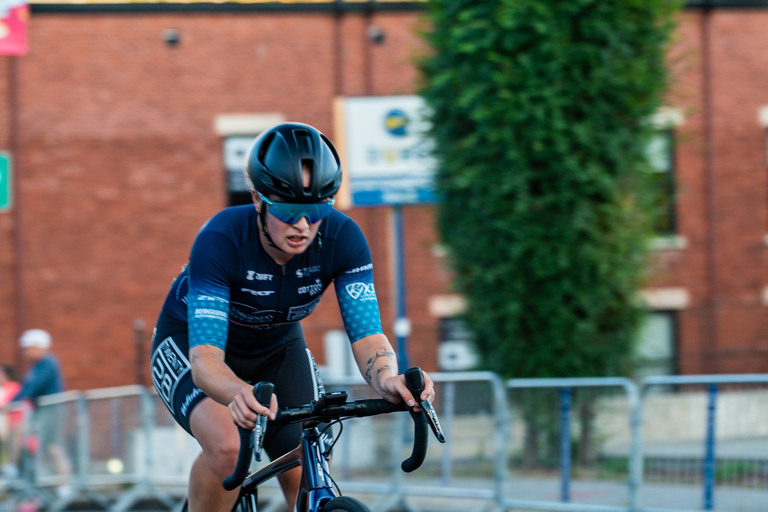 A cyclist riding a black bike on a city street, wearing a black helmet, blue sunglasses, and a blue cycling uniform with logos. In the background, there is a brick building, some railings, and blurred pedestrians.