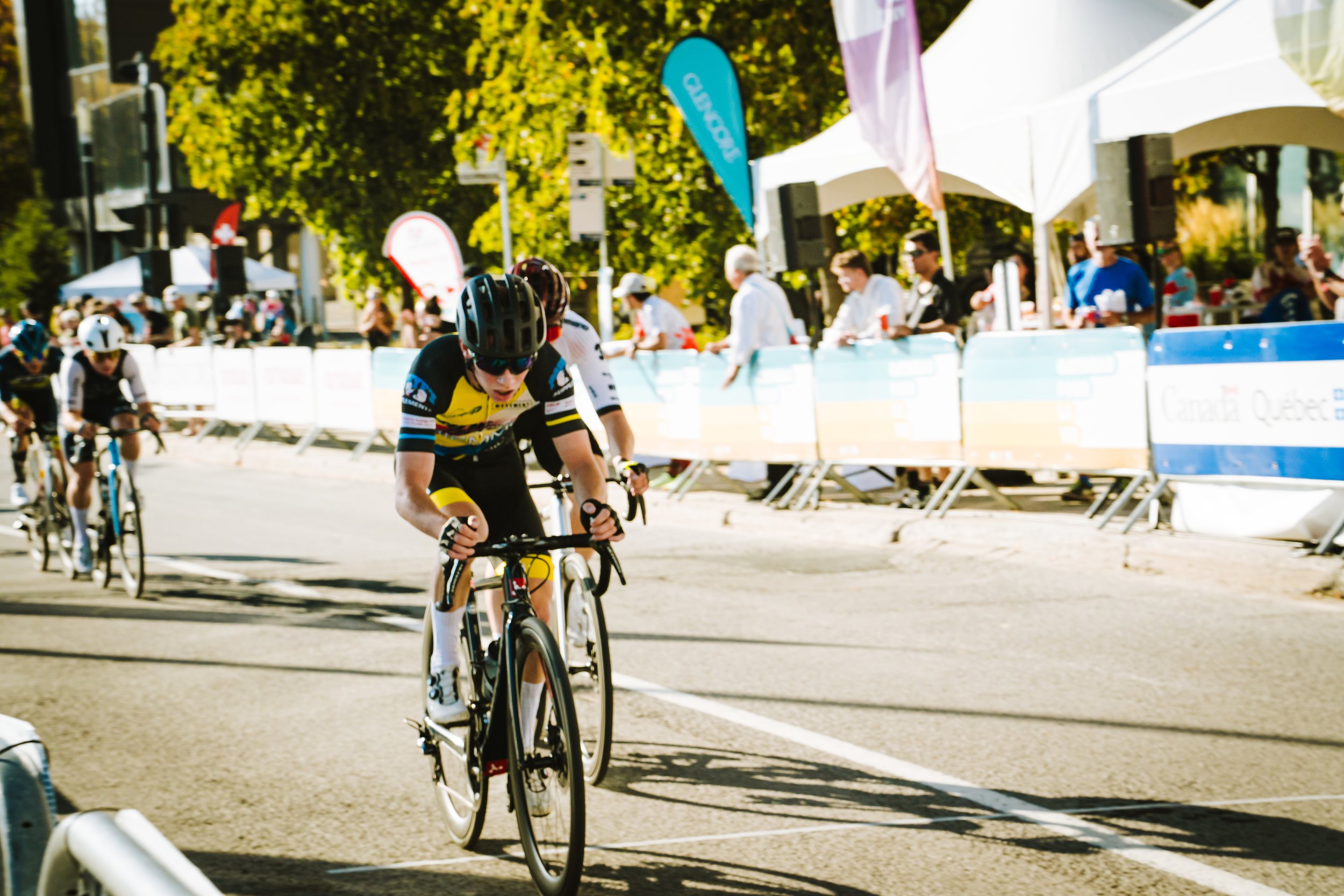 Cyclists race in a biking event on a city street lined with spectators and sponsor tents, with trees and sunny weather.