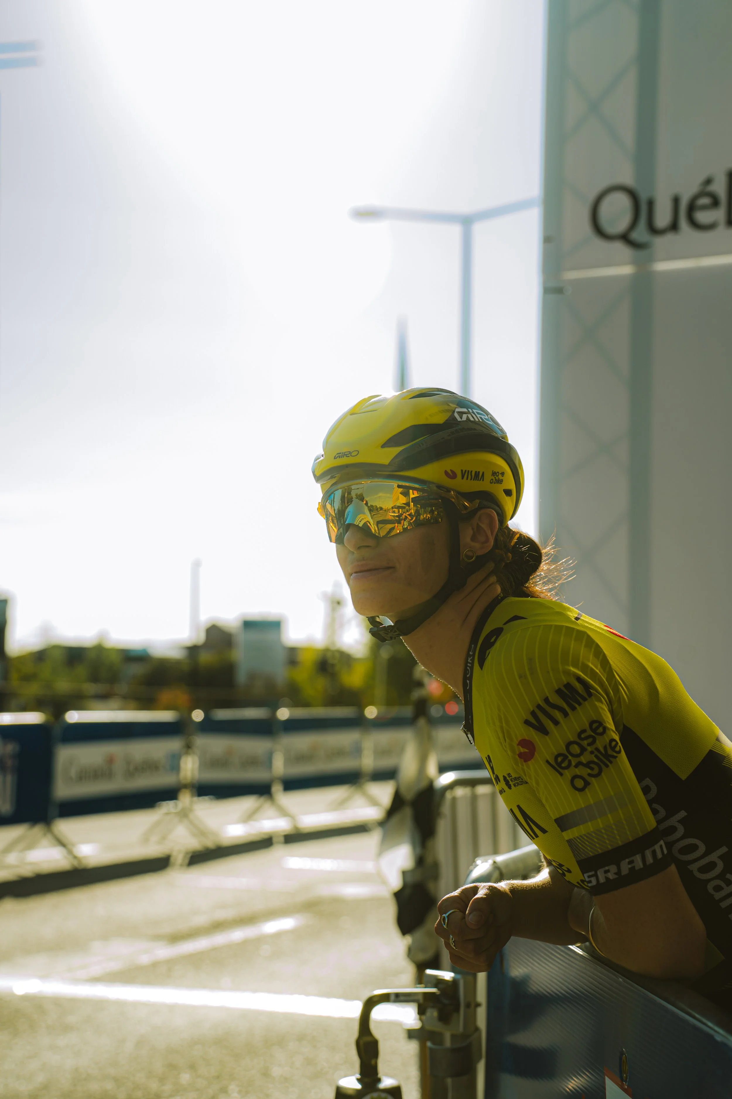 A female cyclist in yellow gear and helmet leaning on a barrier at a race, with a bright sky and some blurred cityscape in the background.