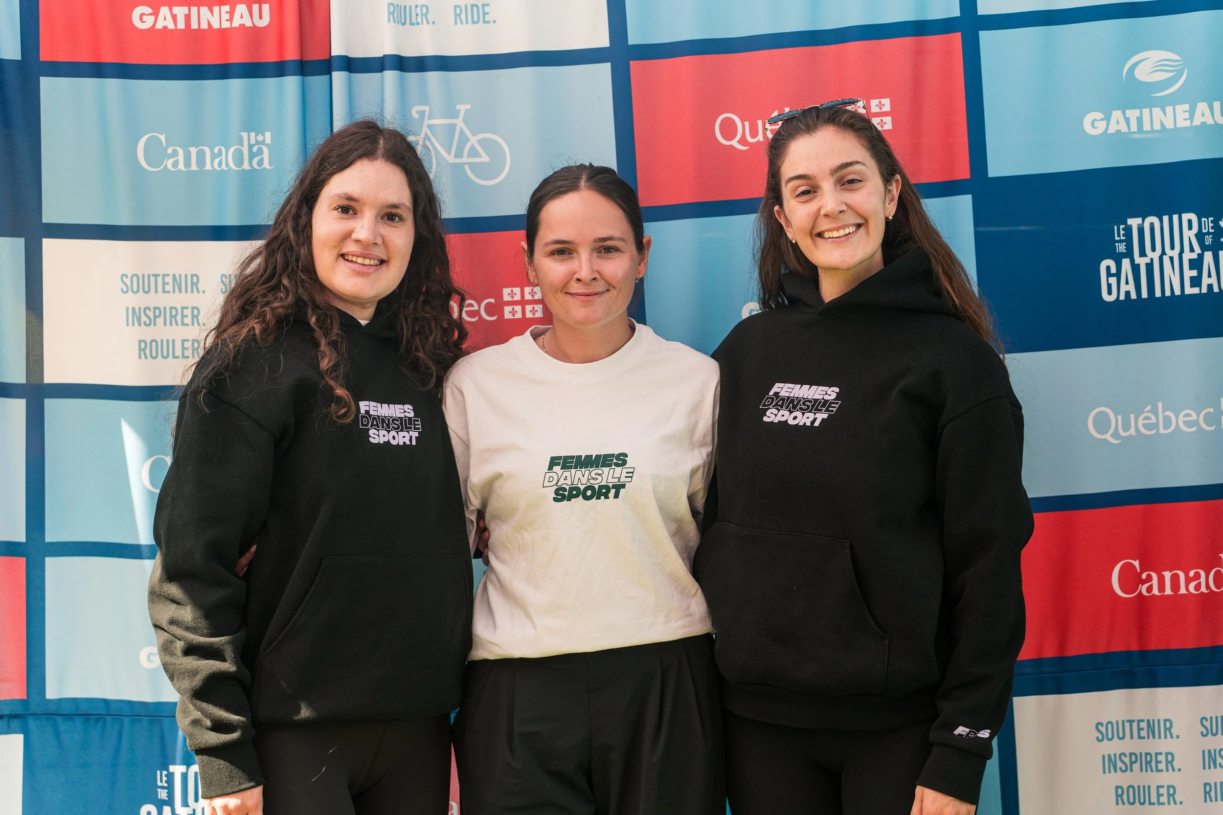 Three women standing together in front of a Canadian-themed backdrop, wearing matching sweatshirts with 'Femmes Dans Le Sport' printed on them, smiling at the camera.