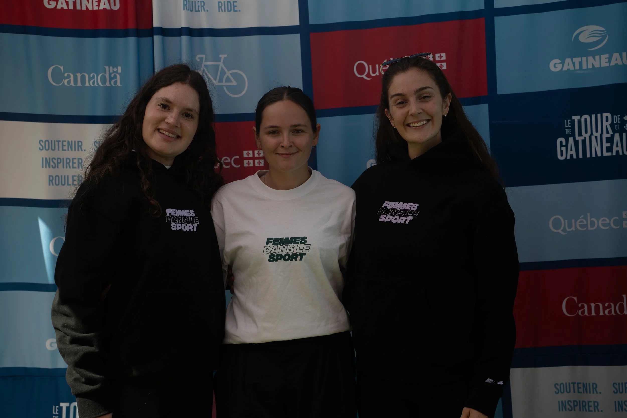 Three women standing together in front of a blue and red promotional backdrop, wearing hoodies and t-shirts with the text 'FEMMES DANS LE SPORT' in French.