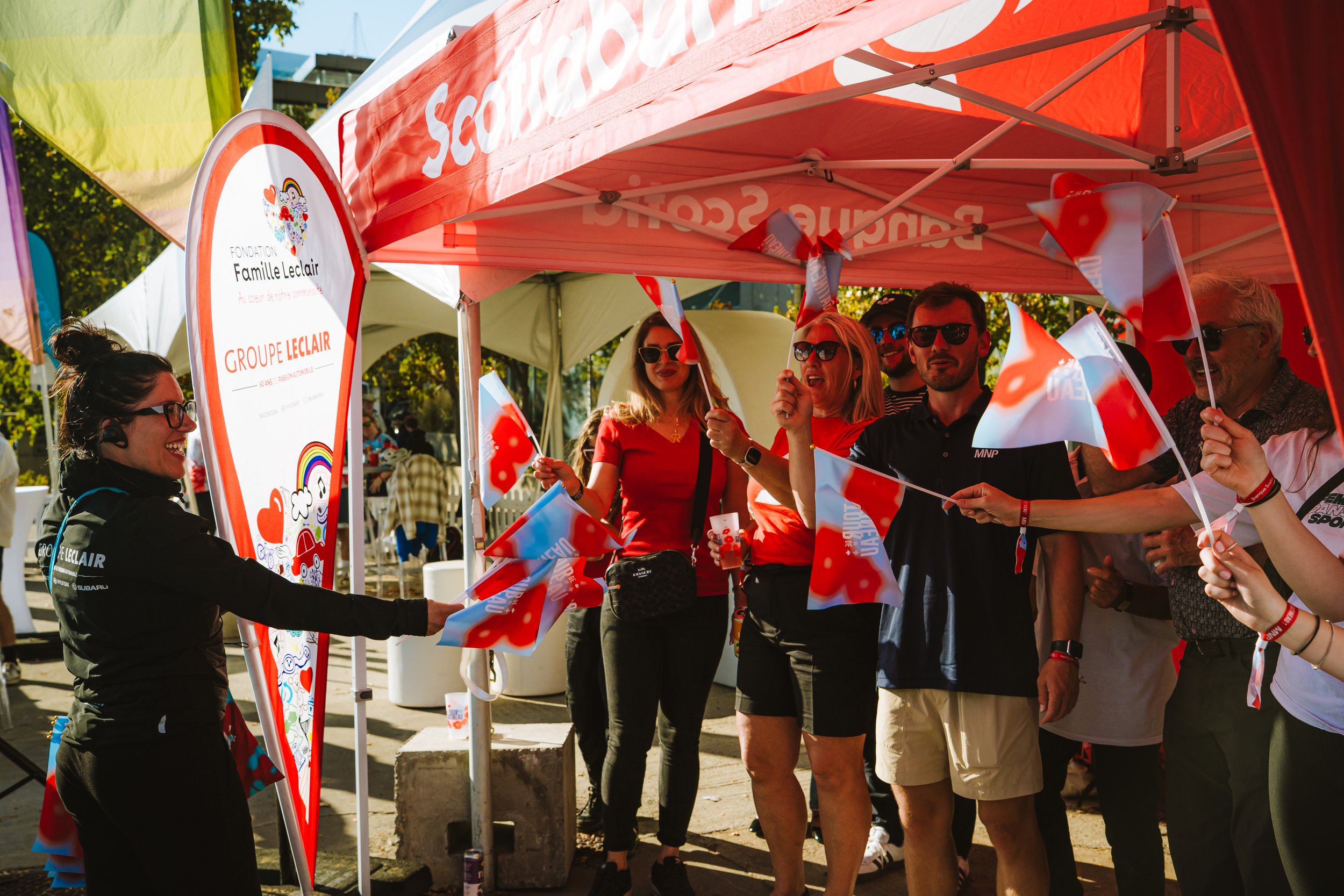 Group of people at an outdoor event under a red tent, holding small flags, wearing sunglasses, and smiling.