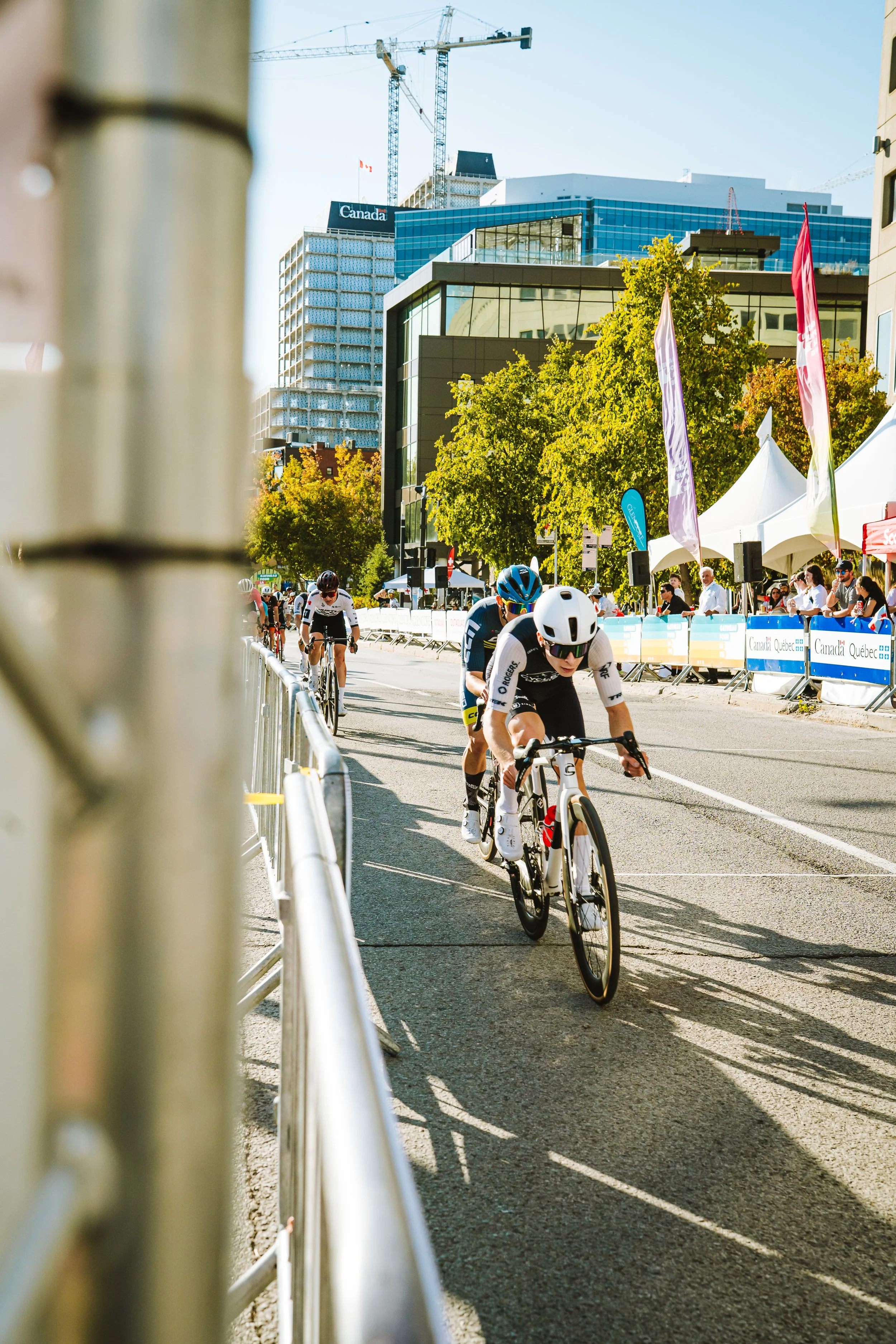 Cyclists participating in a race on a city street, with spectators and tents on the sidewalk, tall modern buildings and construction cranes in the background, and trees lining the street.