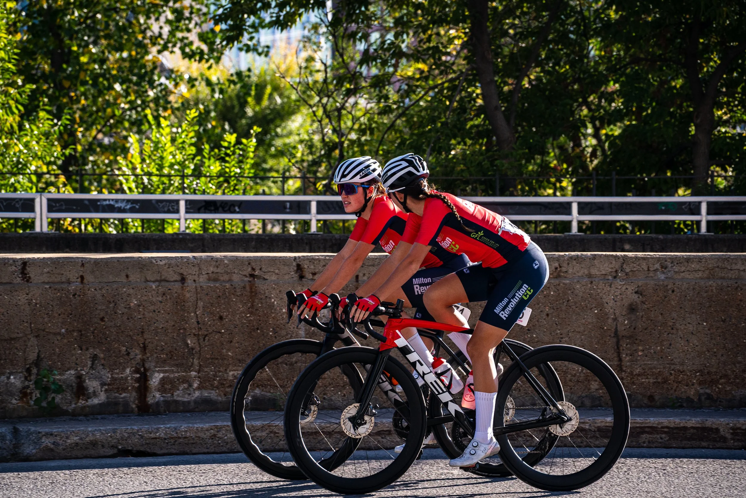 Two female cyclists wearing red jerseys, black shorts, and helmets ride side by side on a paved street with a concrete wall and green trees in the background.
