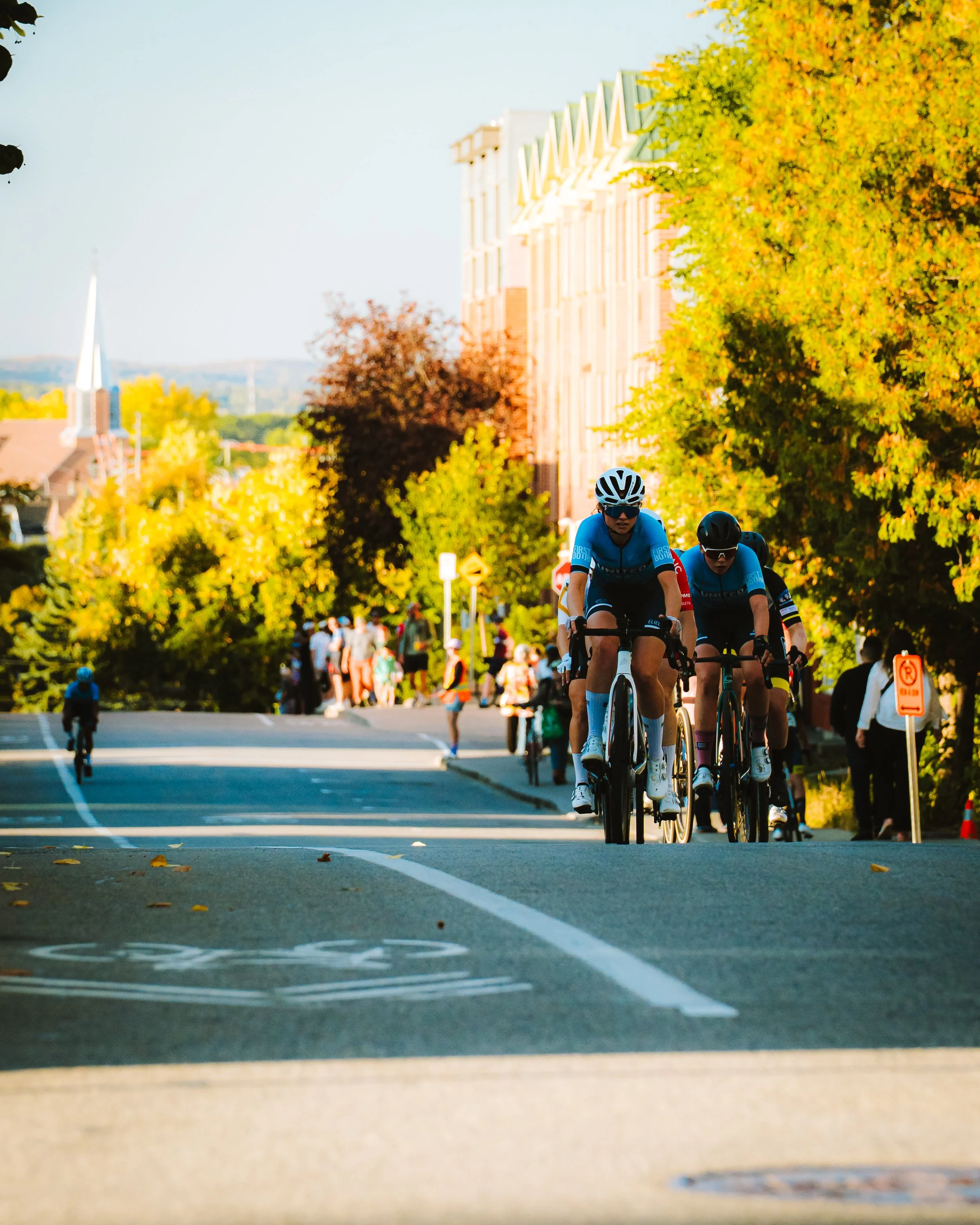Two cyclists riding on a city street, lined with trees and buildings, during daytime with a crowd of pedestrians in the background.