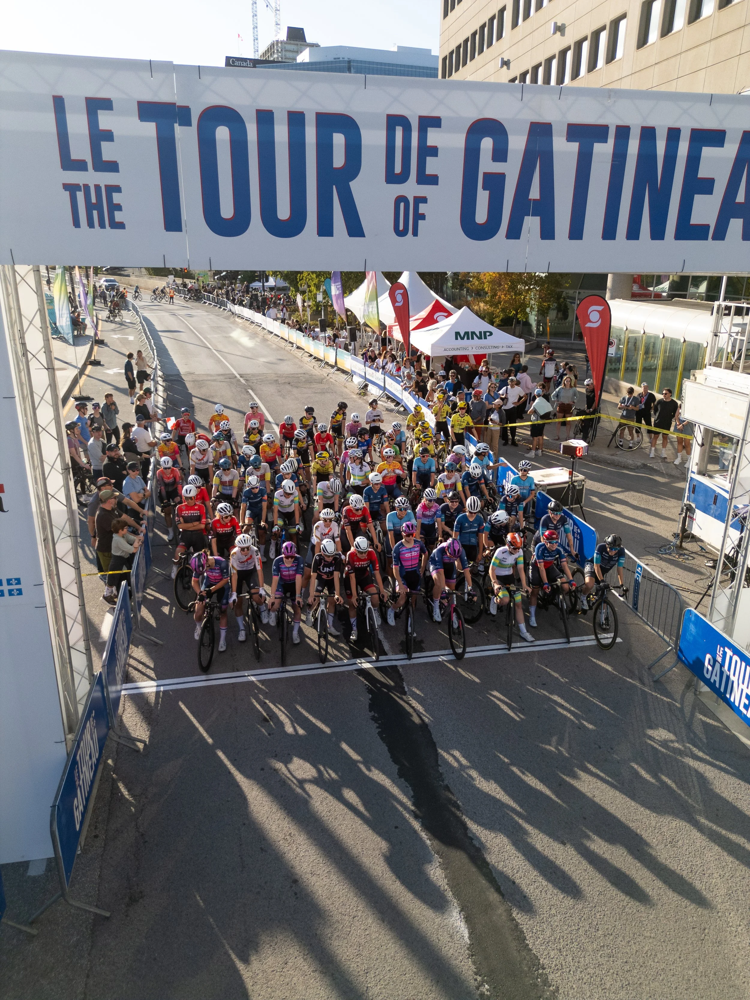 Cyclists at the starting line of the Tour de Gatineau race on a city street, with a large banner overhead and spectators on the sidewalk.