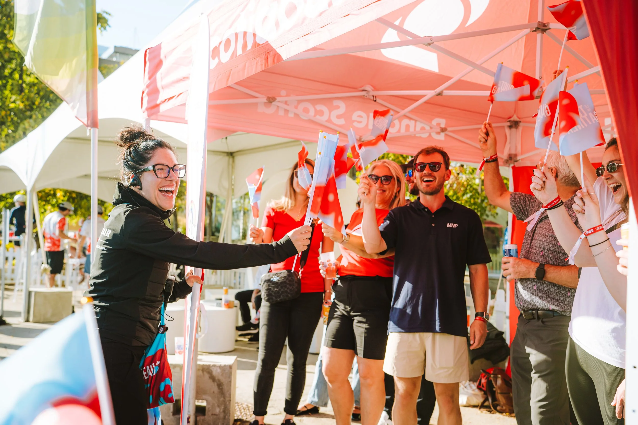 Group of people gathered under a red tent at an outdoor event, waving small flags and smiling.