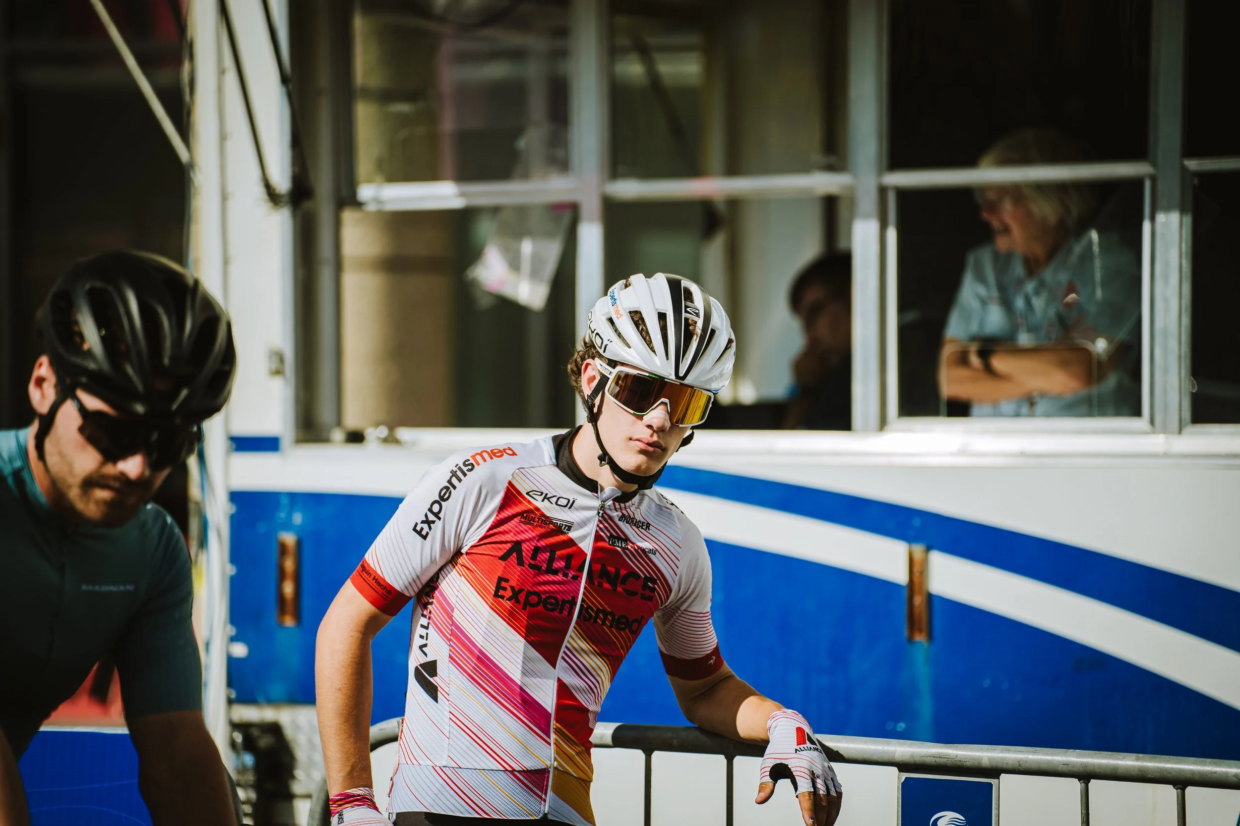 A cyclist in a white and red jersey, wearing a white helmet and tinted glasses, standing beside a barrier. Behind him, there are other people and a building with windows.