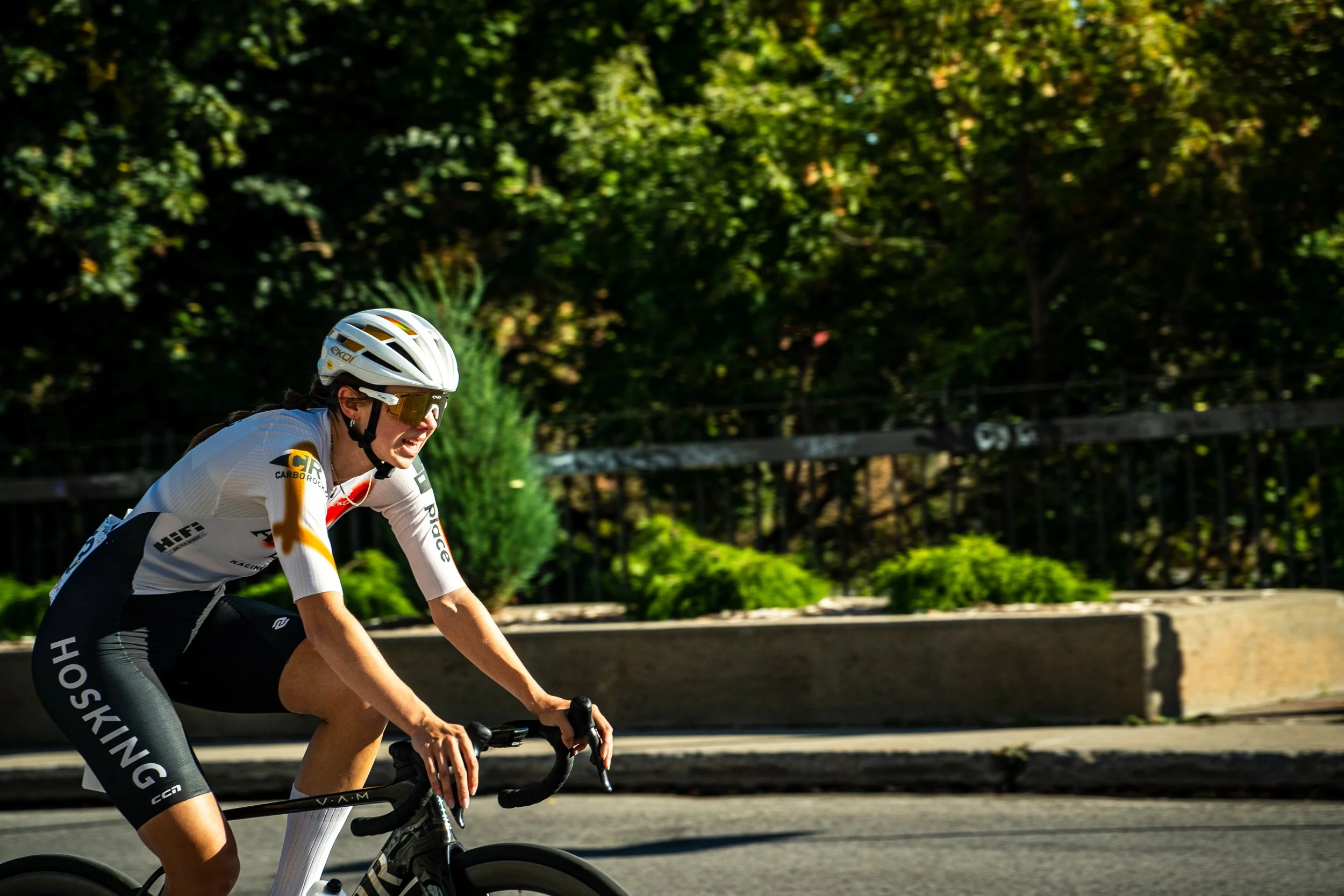 A female cyclist wearing a white helmet, sunglasses, and a racing uniform riding a black road bike on a sunny street with trees and bushes in the background.