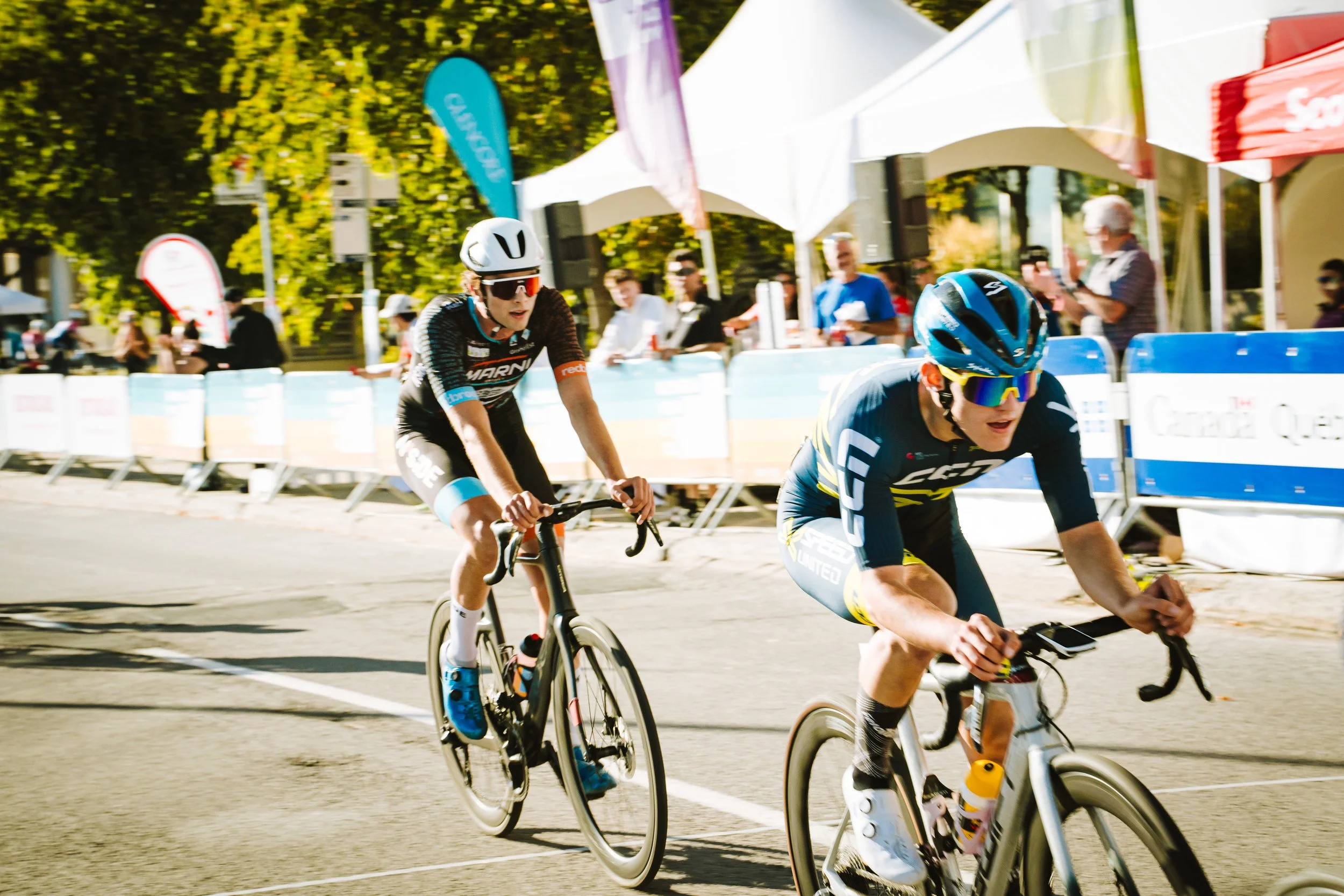Two competitive cyclists race on a city street during a cycling event. The cyclist in front wears a blue and yellow jersey and a blue helmet, while the cyclist behind wears a black jersey with blue accents and a white helmet. Spectators line the stre