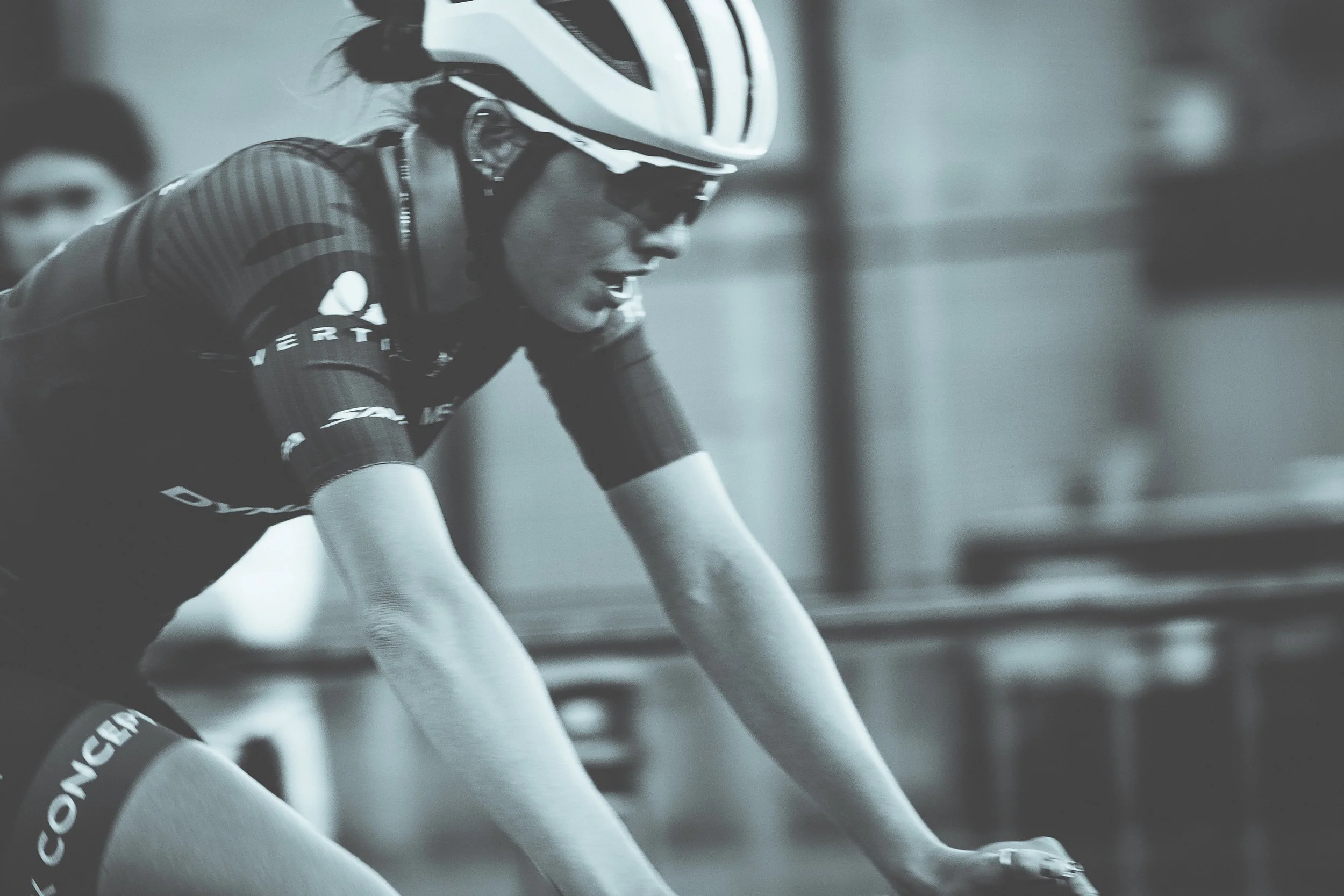 A female cyclist wearing a helmet, sunglasses, and a cycling jersey, leaning forward on a stationary bike, with a blurred background.