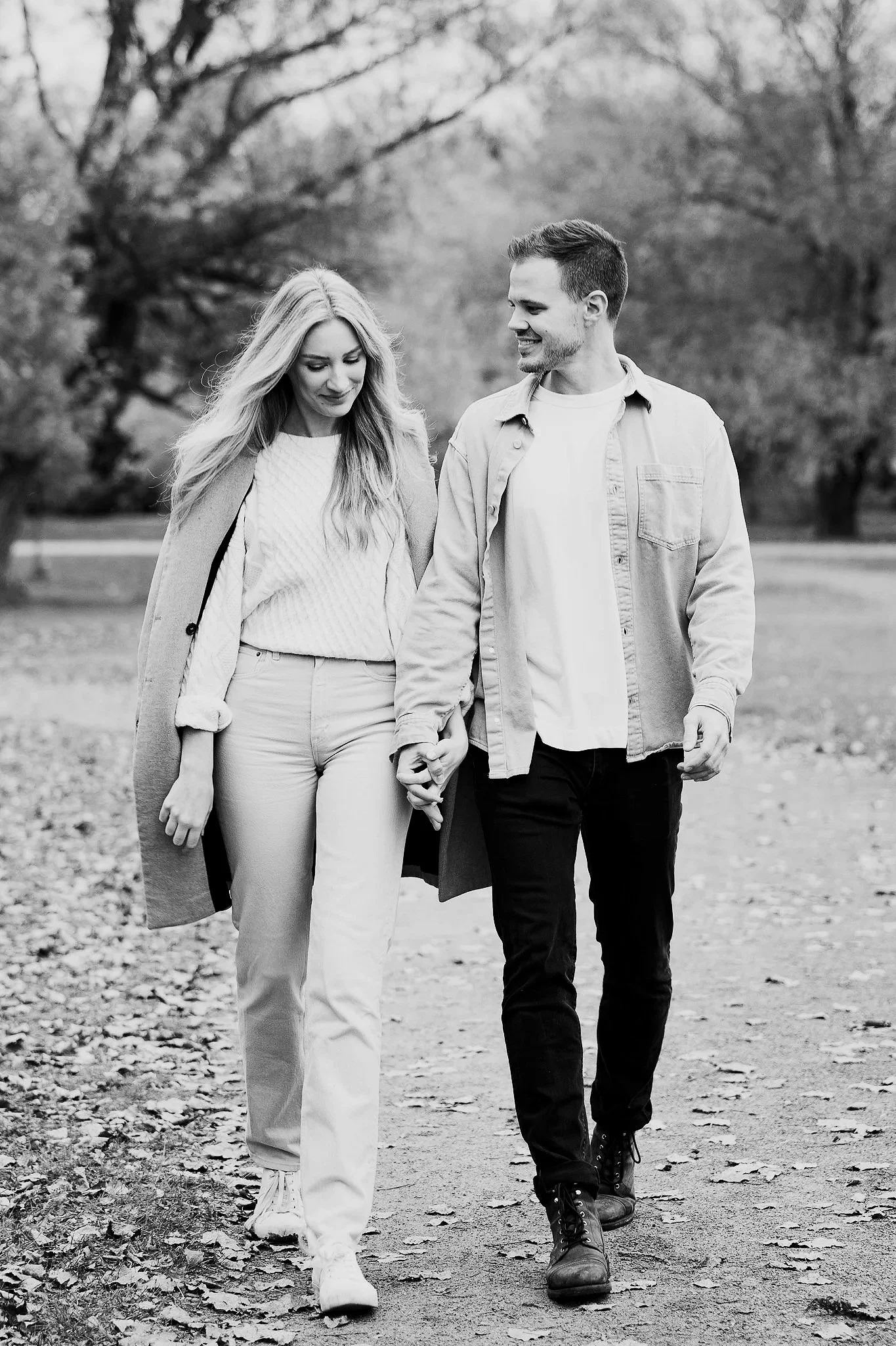 A young couple walking hand-in-hand through a park during autumn, surrounded by trees with fallen leaves.