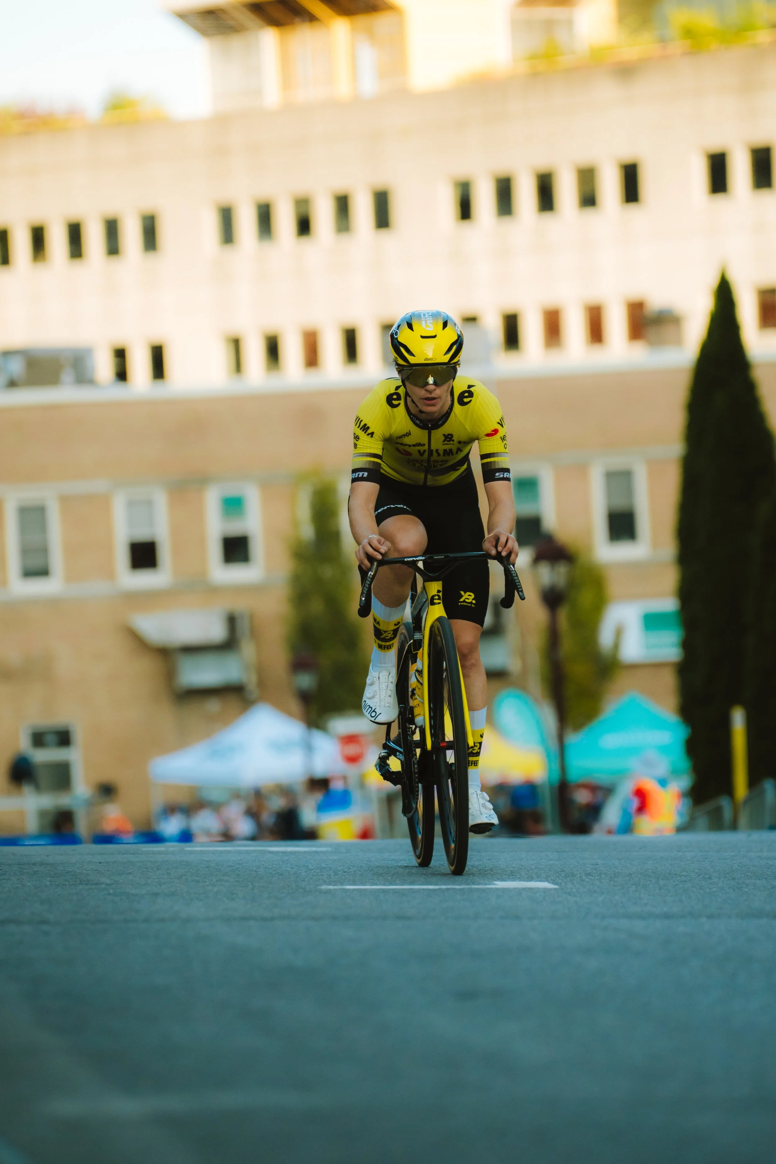 A cyclist in yellow racing gear riding a black and yellow bike on a city street with buildings and tents in the background.