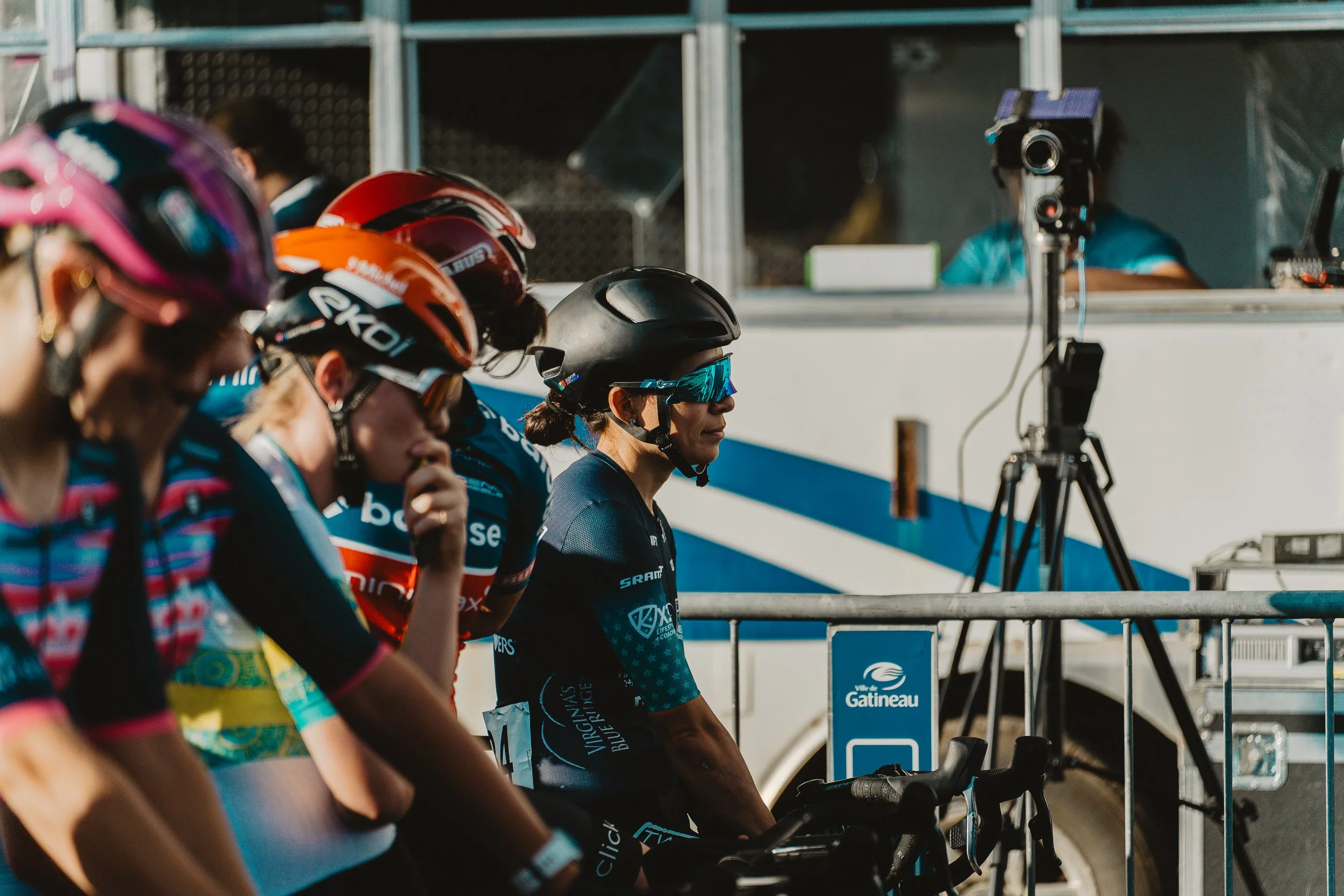 Cyclists wearing helmets and sunglasses, preparing at a starting line during a race, with a vehicle and camera setup in the background.