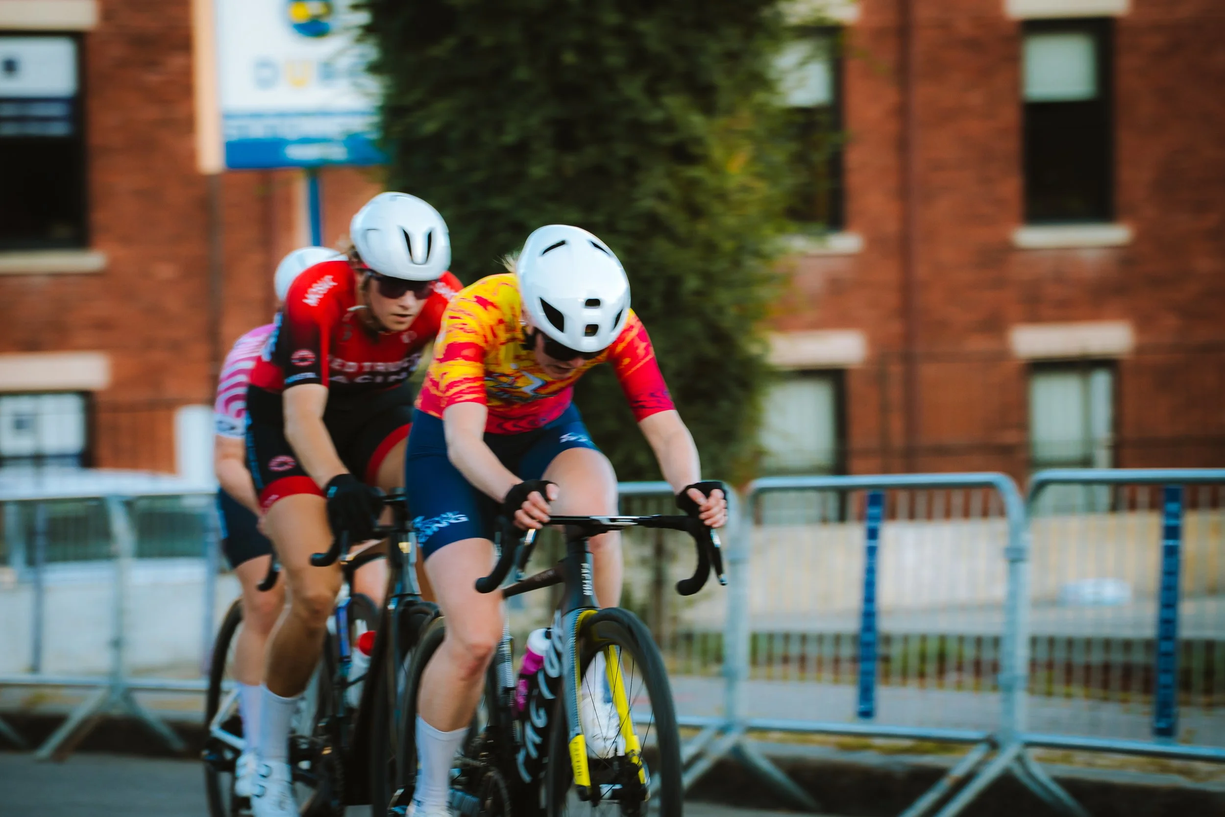 Cyclists racing in a competitive event, wearing helmets and team jerseys, on a city street with barriers and a brick building in the background.