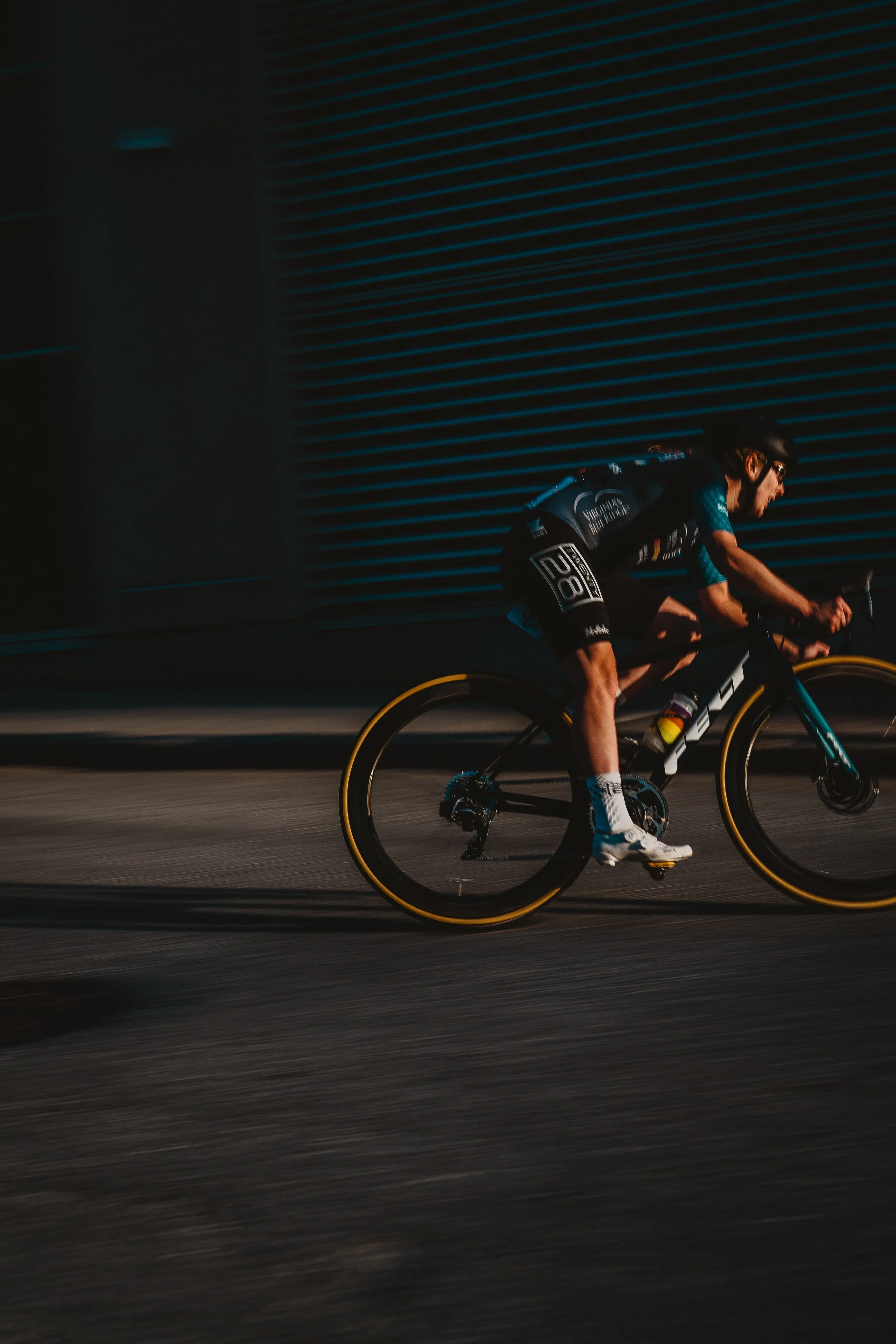 A cyclist wearing a helmet and sports gear riding a black and teal mountain bike with yellow accents on the tires, in motion along a dark, urban street.