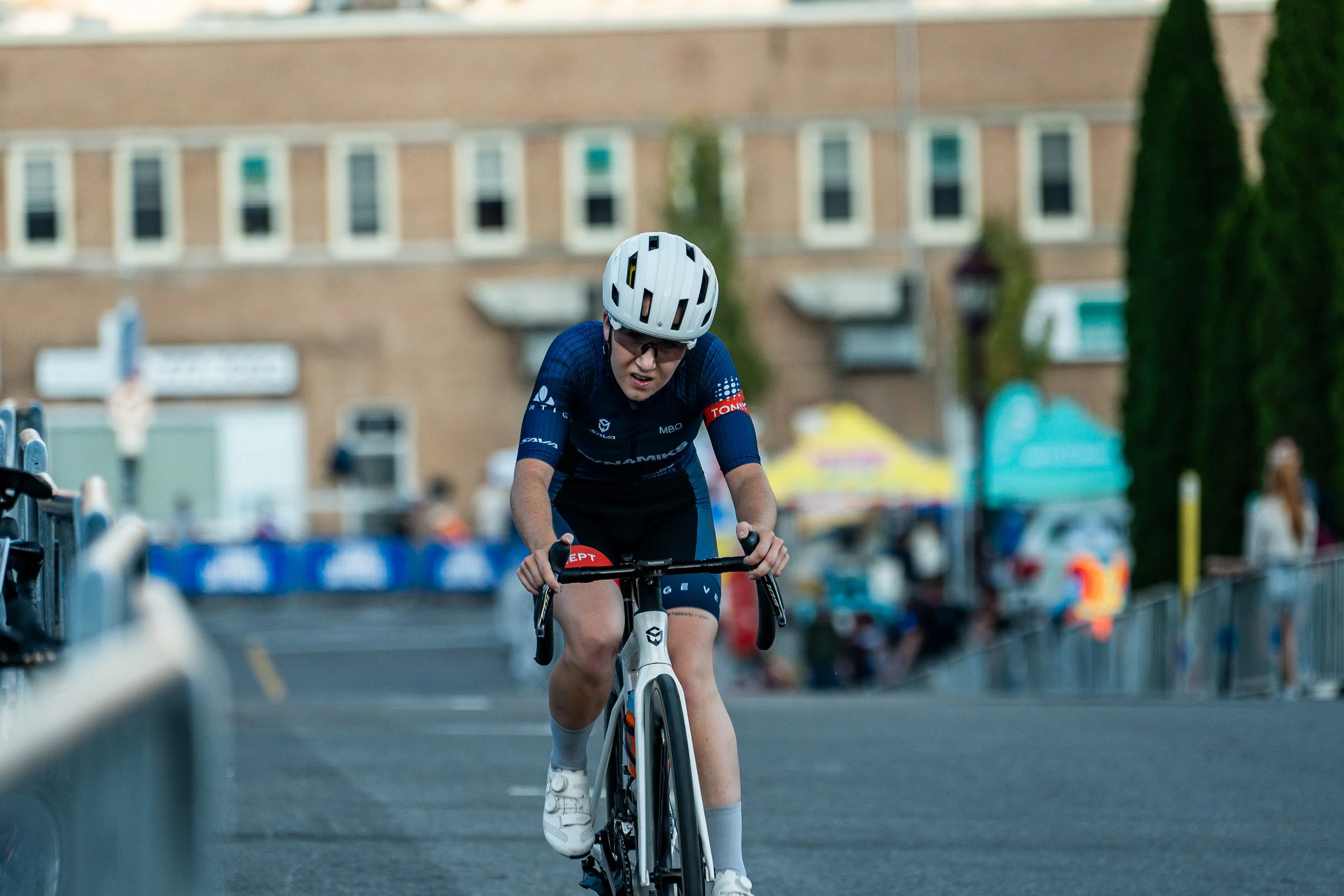A cyclist wearing a white helmet, sunglasses, and a dark blue cycling jersey with red and white accents riding on a city street during a race.