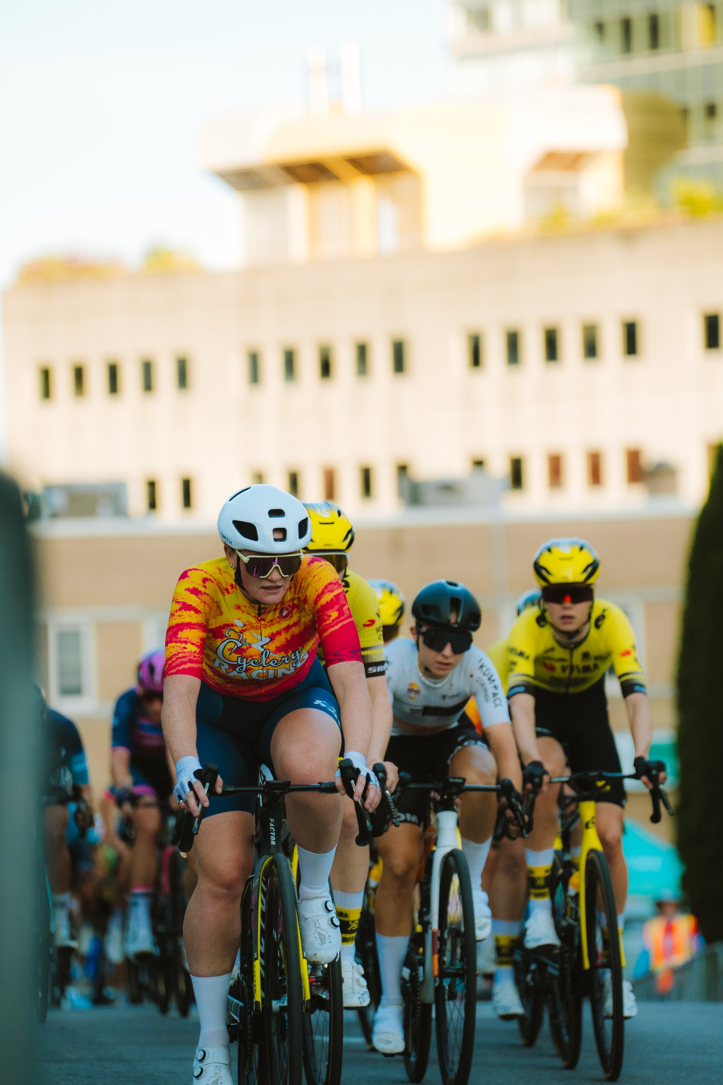 Cyclists in a race, wearing colorful jerseys and helmets, riding on city streets with buildings in the background.